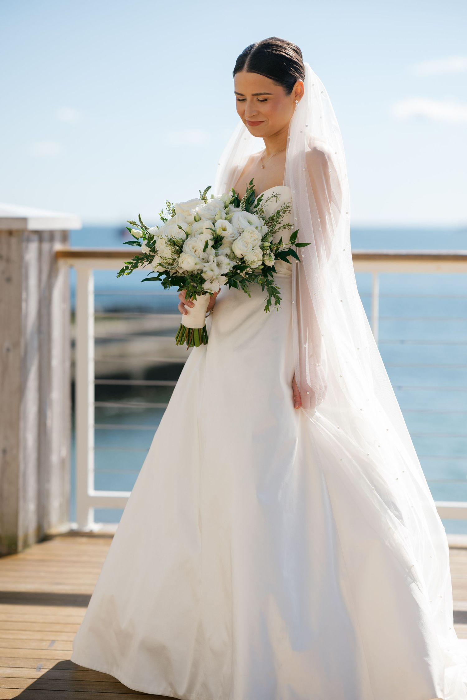 Bride in white gown and veil holds bouquet of white flowers on seaside deck under blue sky.