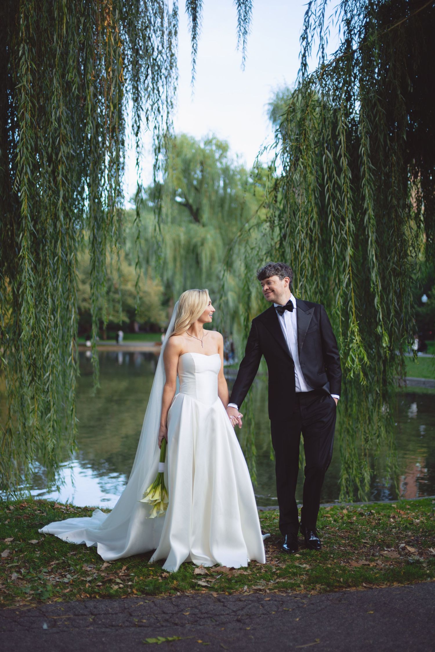Bride and groom hold hands under weeping willow tree by pond, she wears white gown with veil, he wears black tuxedo.