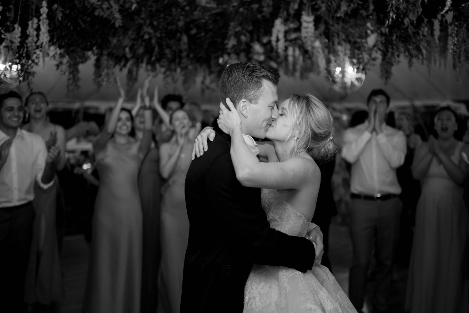 Black and white wedding reception photo showing couple dancing surrounded by guests under twinkling lights.