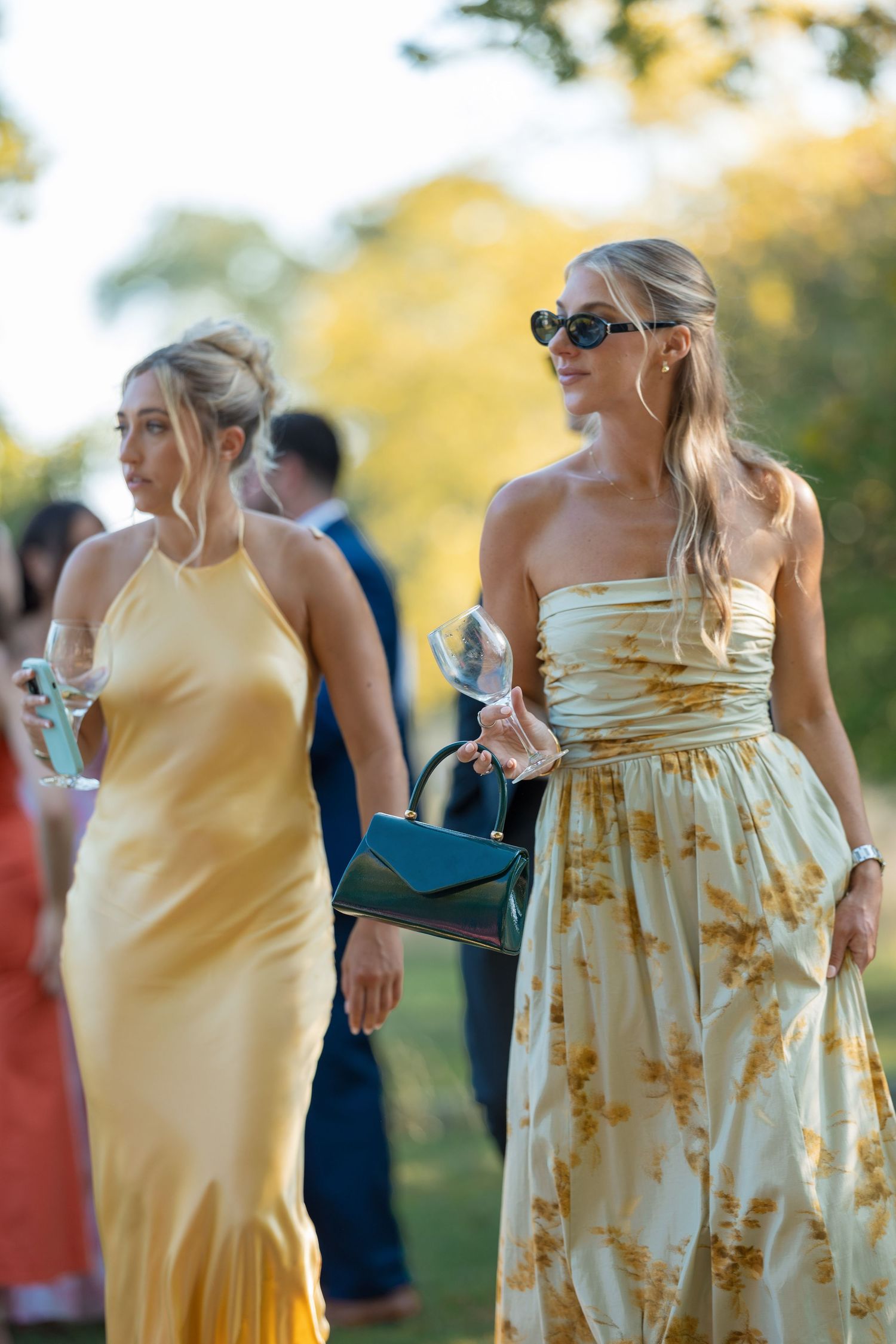Two blonde women in yellow dresses holding champagne glasses at outdoor wedding reception.