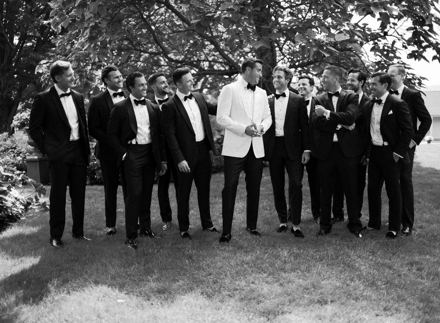 A group of groomsmen in black tuxedos and one in a white dinner jacket pose together outdoors for a formal wedding photo.