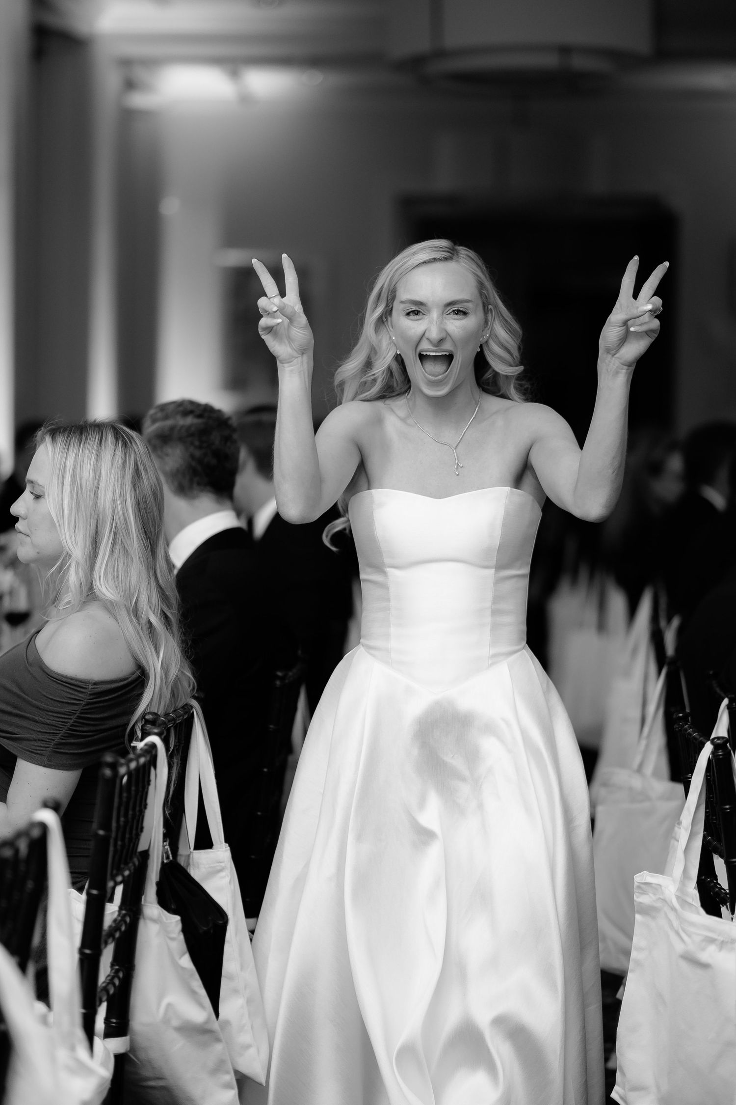 Black and white photo of a joyful bride in a strapless gown making peace signs at her wedding reception.