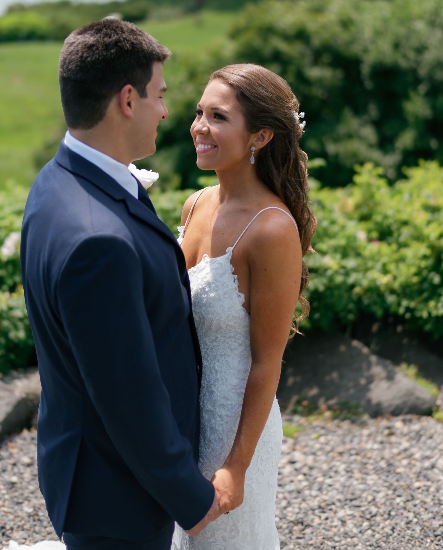 Bride and groom holding hands and smiling at each other outdoors on their wedding day with lush green scenery.