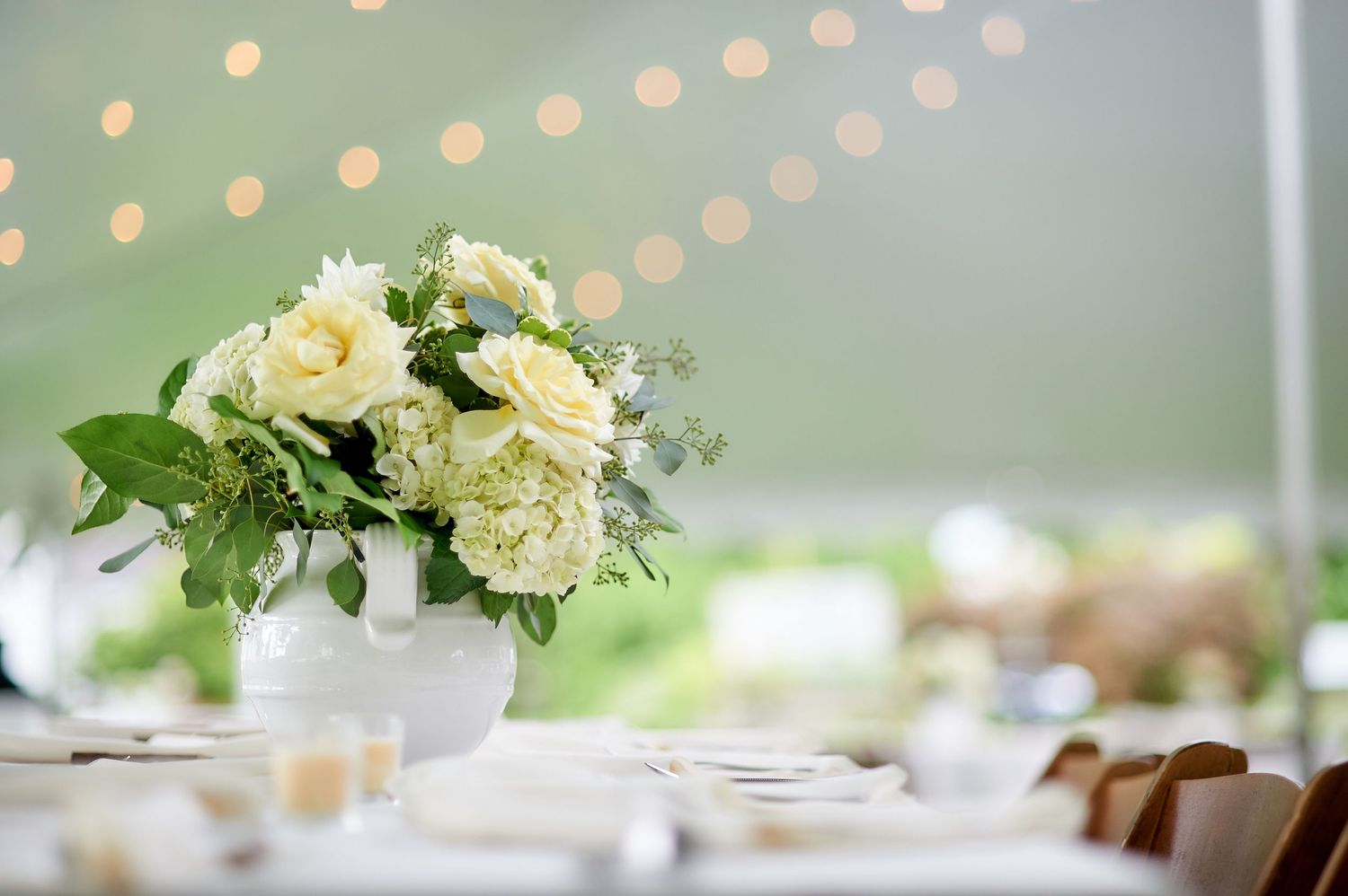 Elegant white floral centerpiece arrangement on wedding reception table with bokeh lights in background.