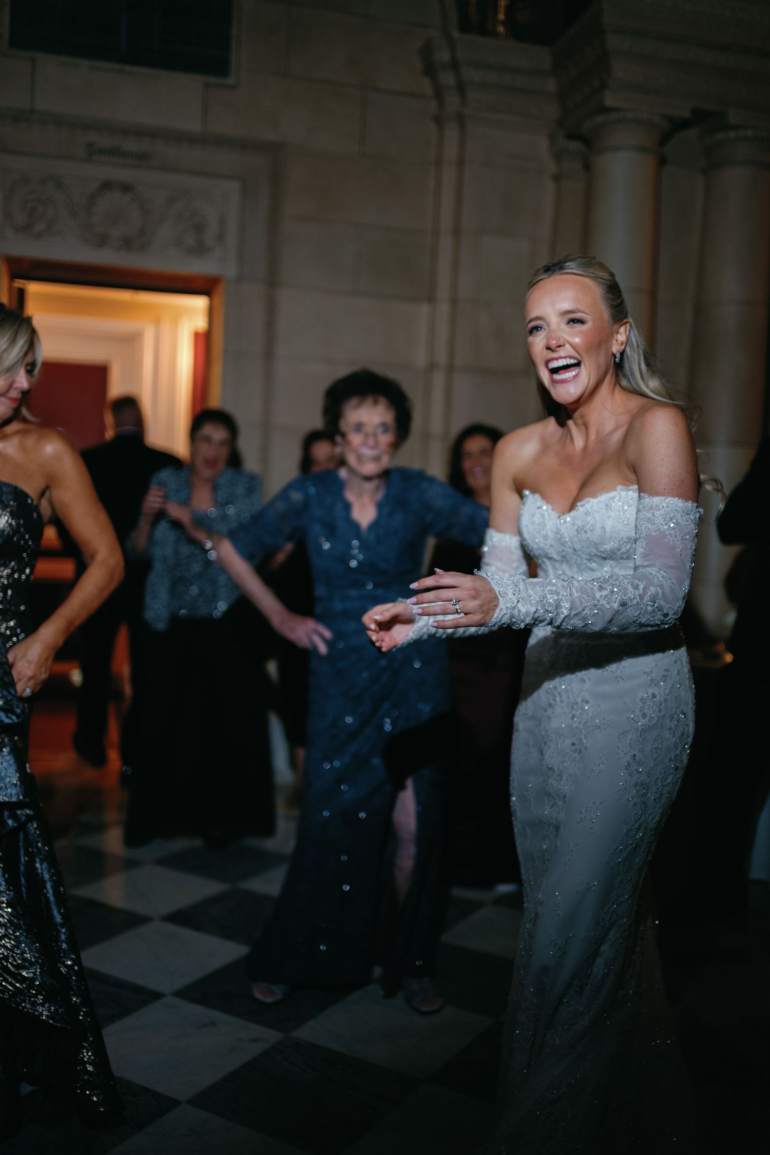 Joyful bride in a fitted white off-shoulder gown laughs while dancing at her wedding reception on a checkered floor.