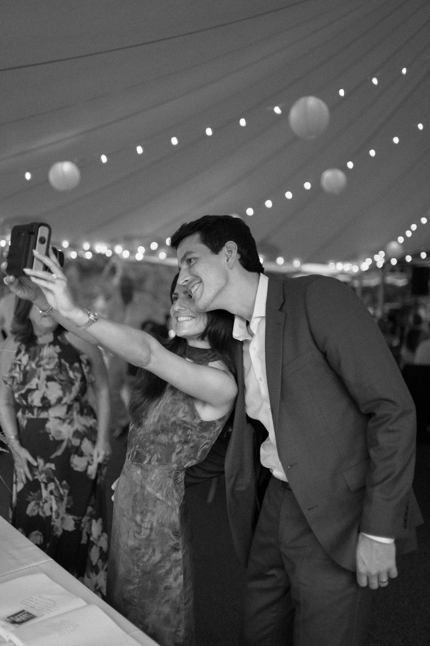 Couple taking a selfie at a wedding reception under string lights in a tent, black and white photo.