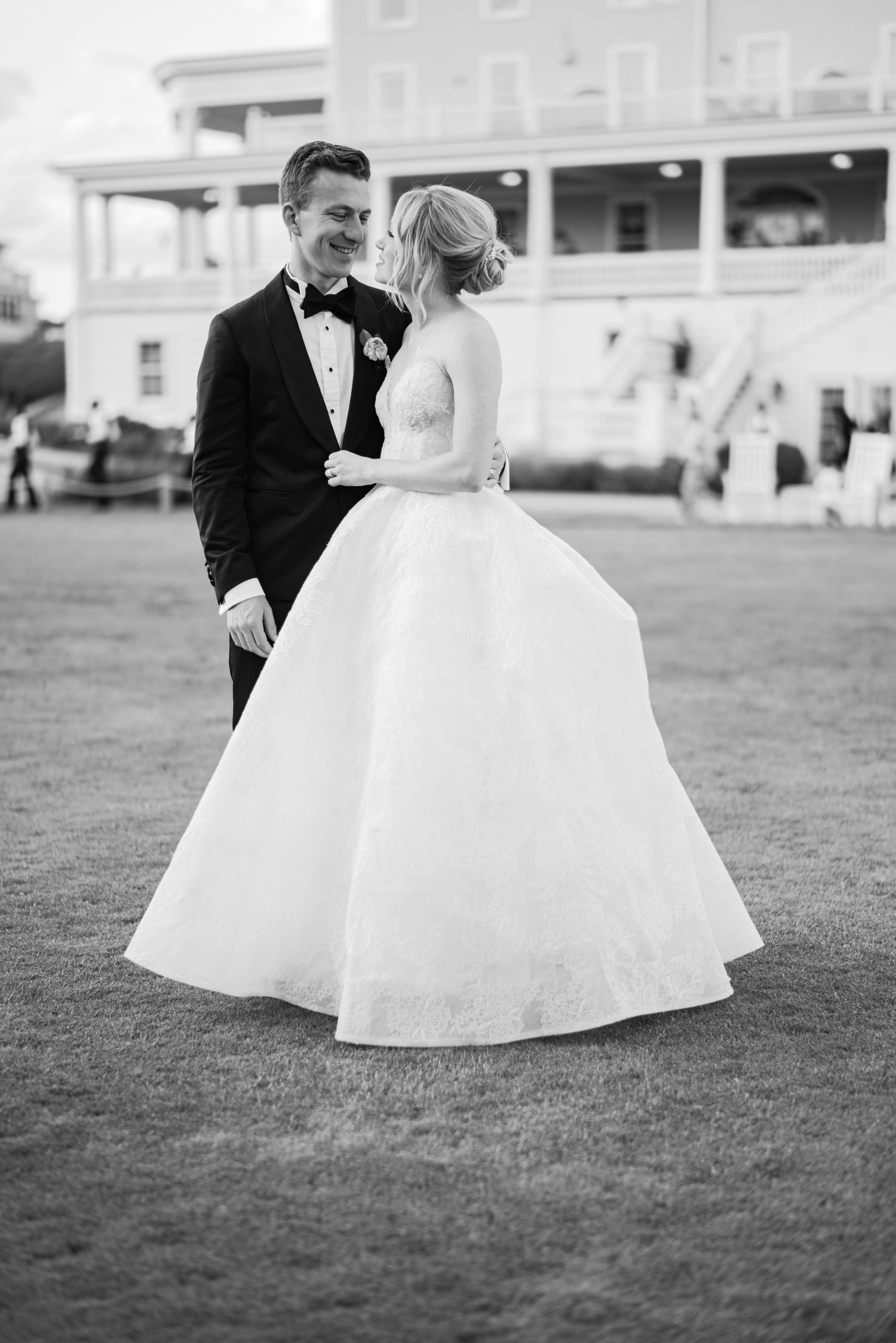 A bride in a flowing white ball gown twirls with her groom on a grassy lawn in black and white photography.