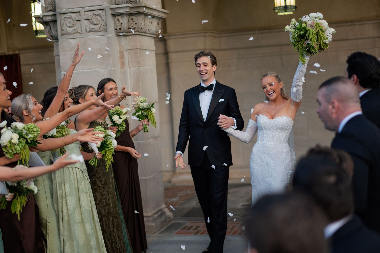 Wedding party celebrates joyfully with confetti and flowers during outdoor ceremony procession.