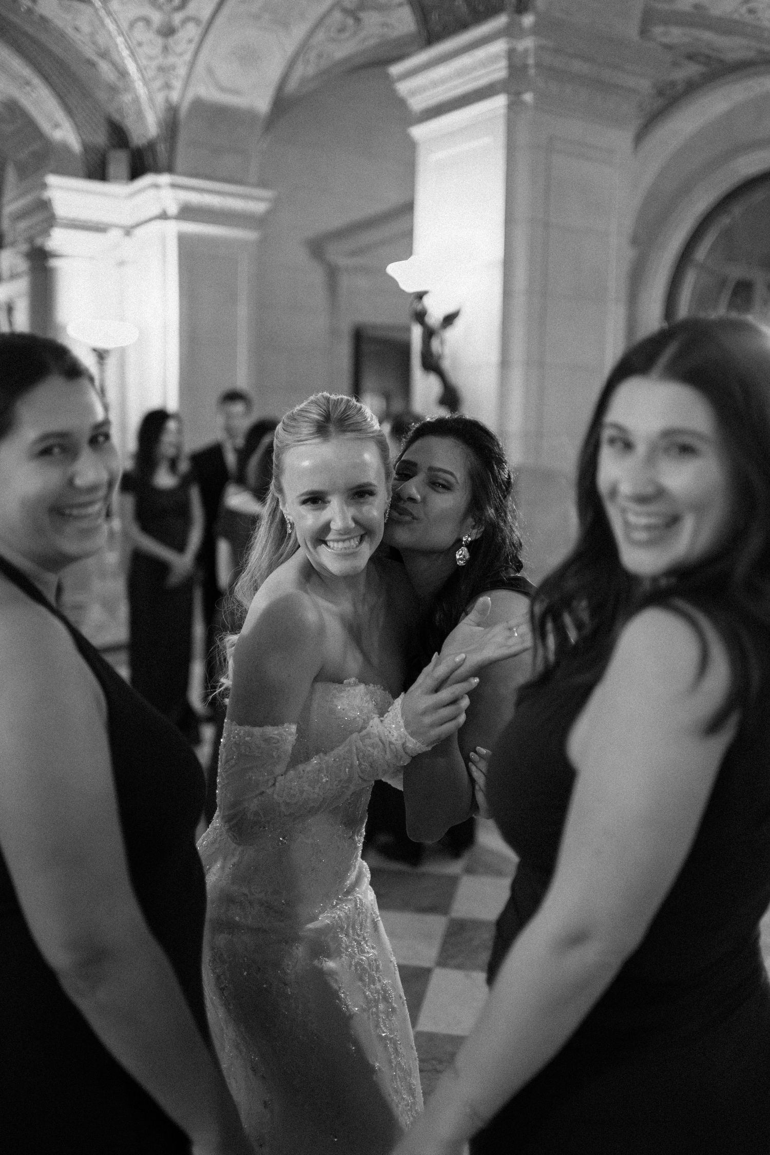 Black and white photo of a bride and friends celebrating and laughing at an elegant reception venue with arched columns.
