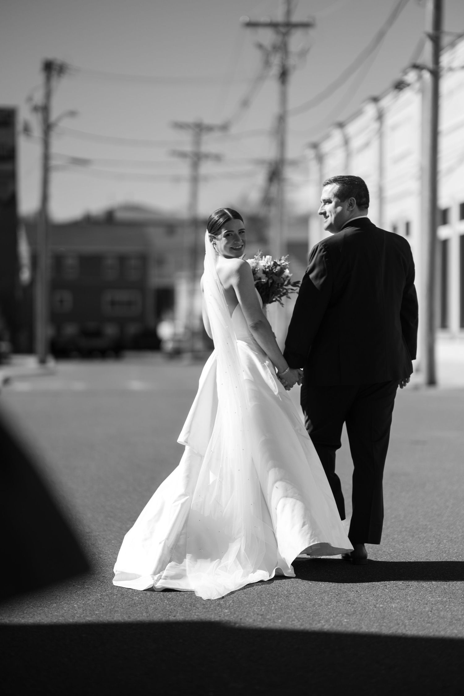 Black and white photo of a bride and groom walking hand in hand down a sunny street, bride holding a bouquet.