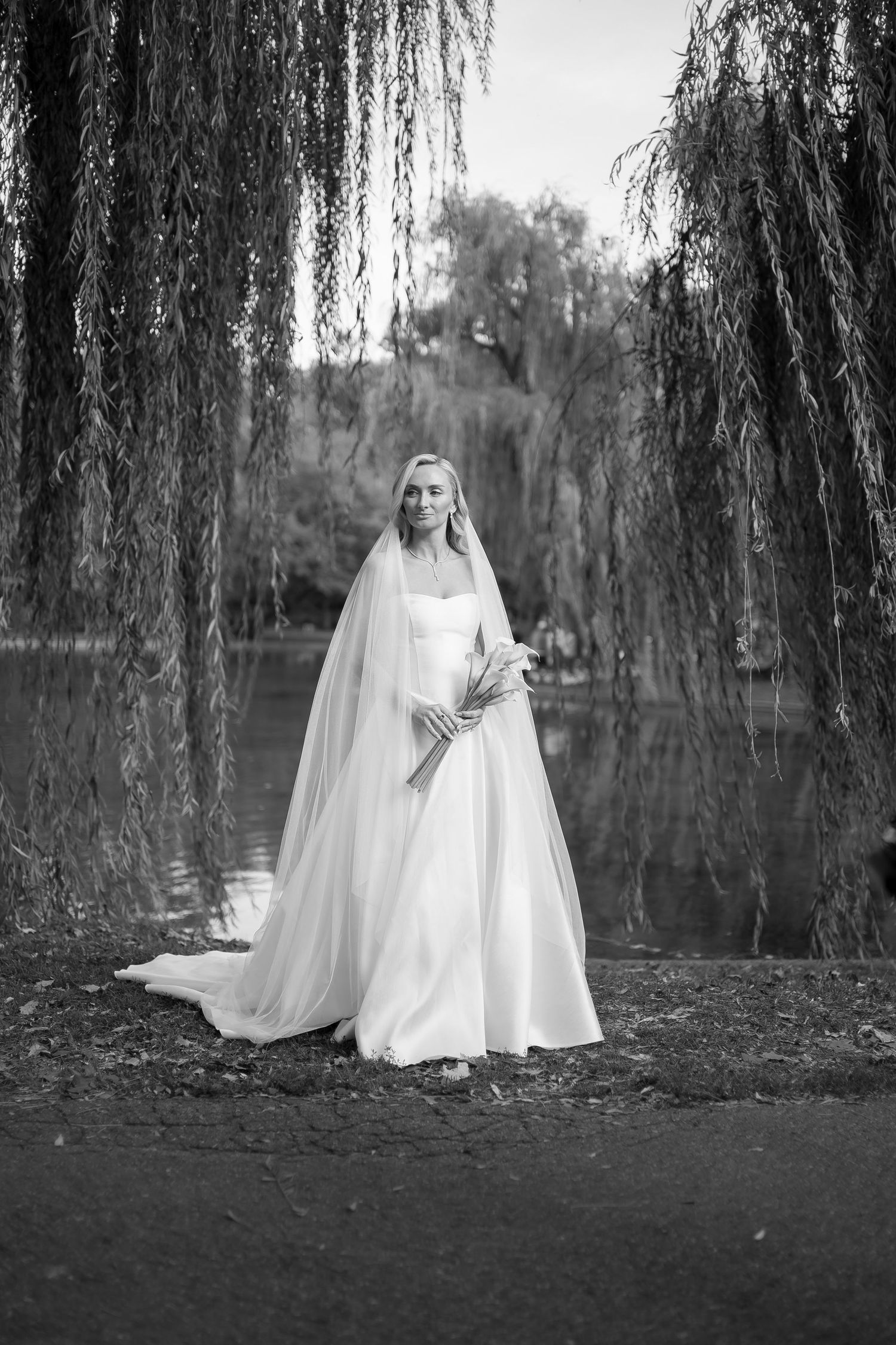 Black and white photo of a bride in a white gown and veil standing beneath a weeping willow tree by a lake.
