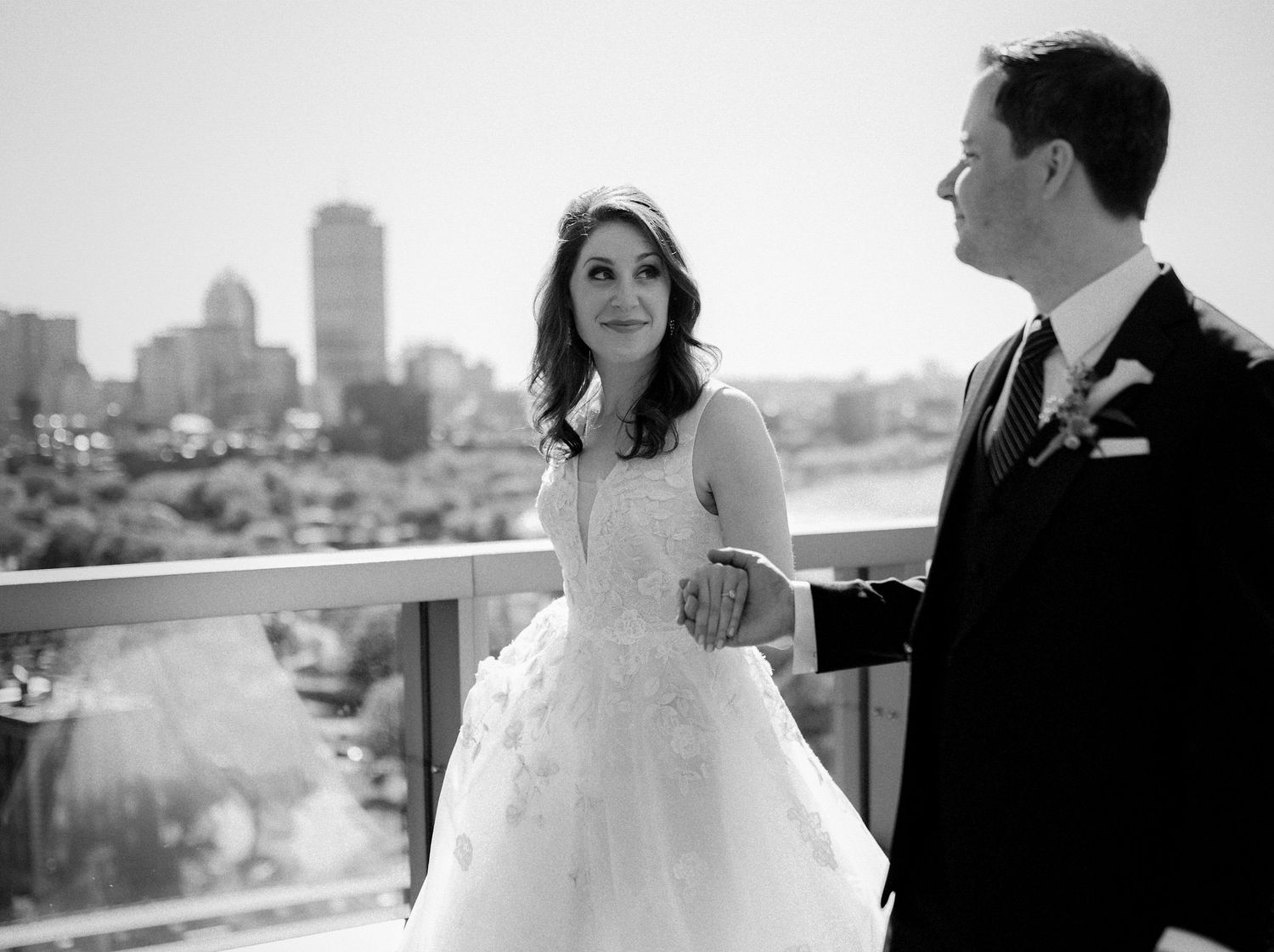 Black and white wedding photo of bride and groom holding hands on a balcony with city skyline backdrop.