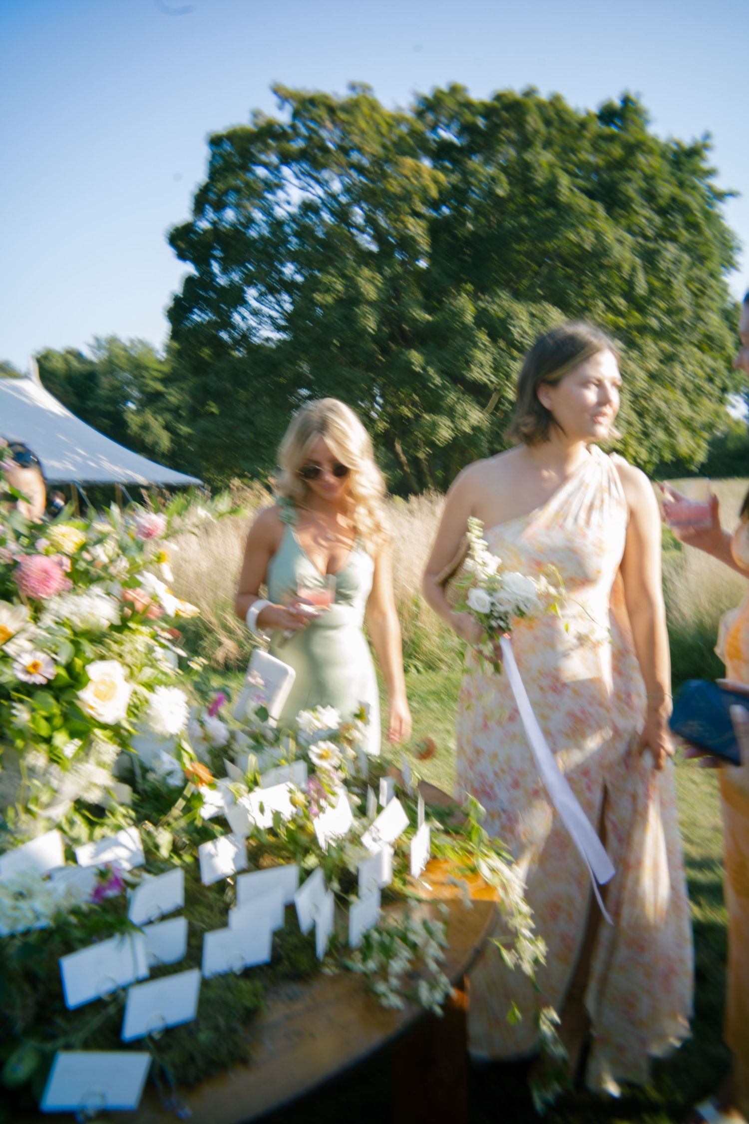 Wedding party members walk past floral displays and place cards in outdoor garden setting.