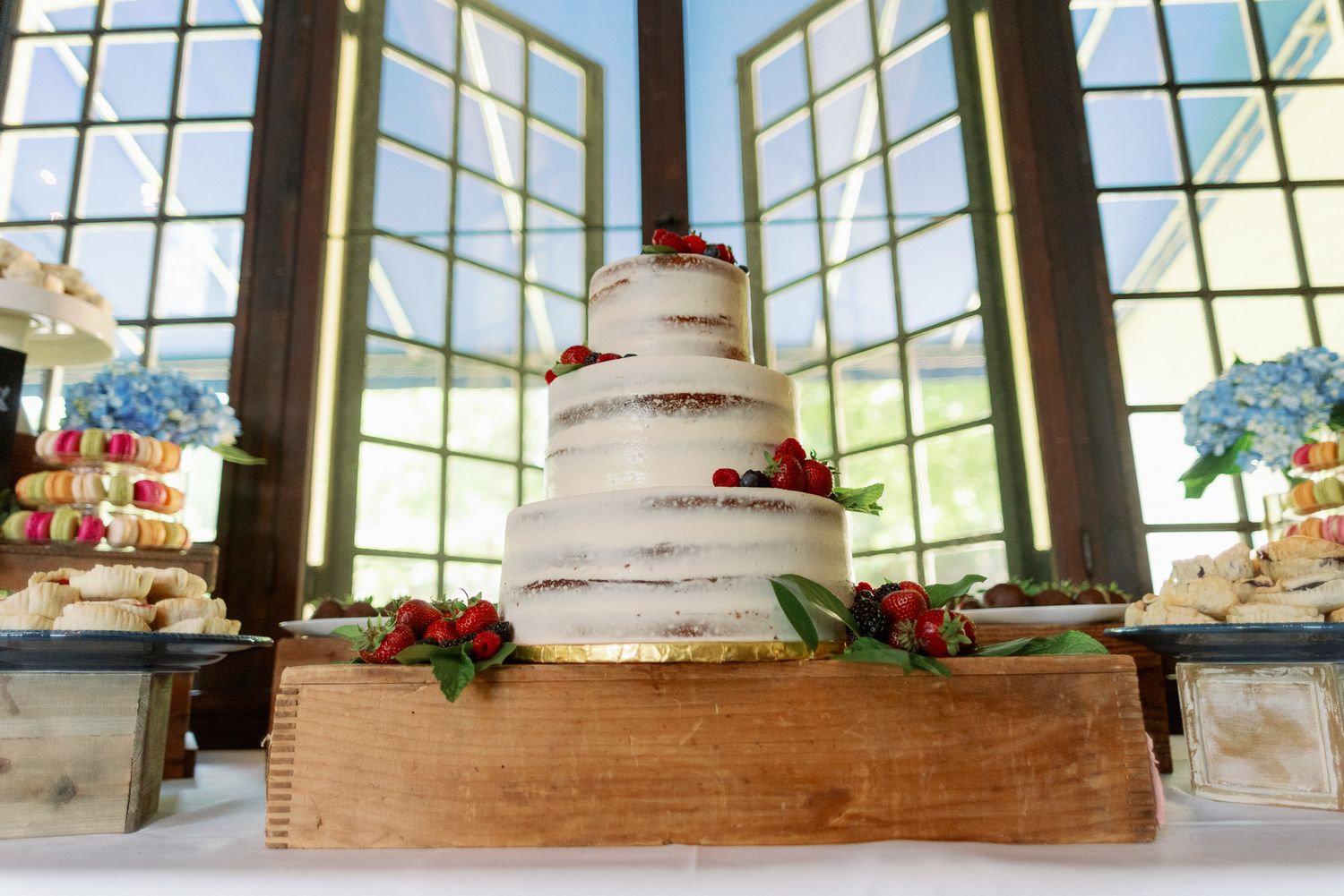 A white tiered wedding cake displayed in front of large windows with floral decorations.