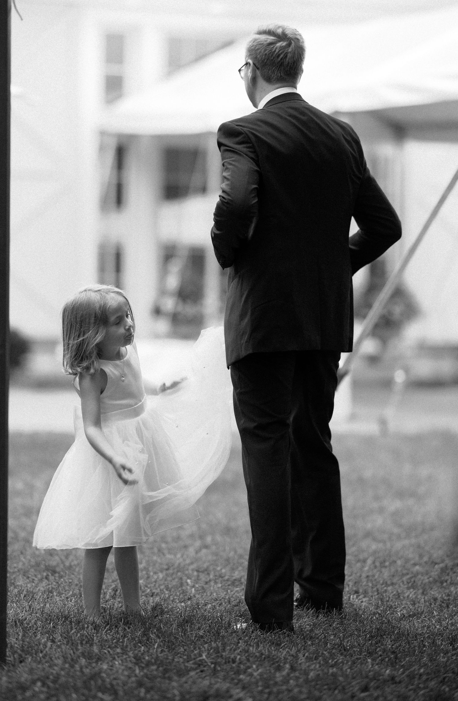 A young girl in a white dress stands on grass looking up at someone in a dark suit viewed from behind.