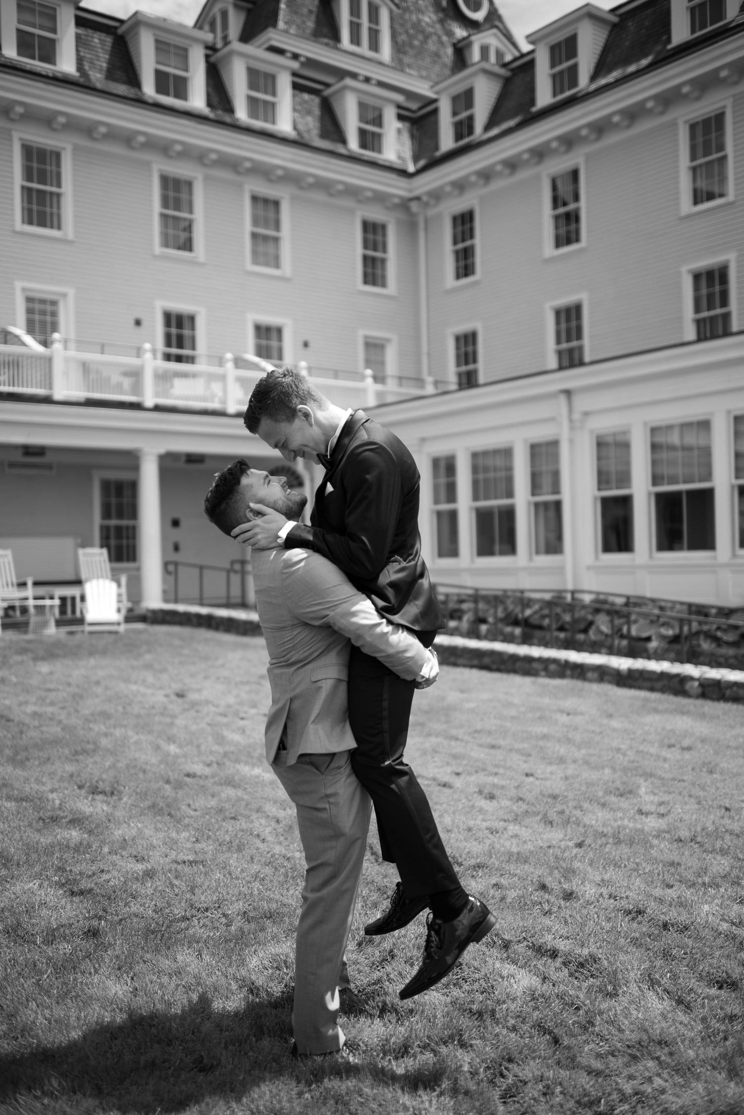 A couple shares a romantic kiss on a grassy lawn in front of a grand historic hotel in black and white.