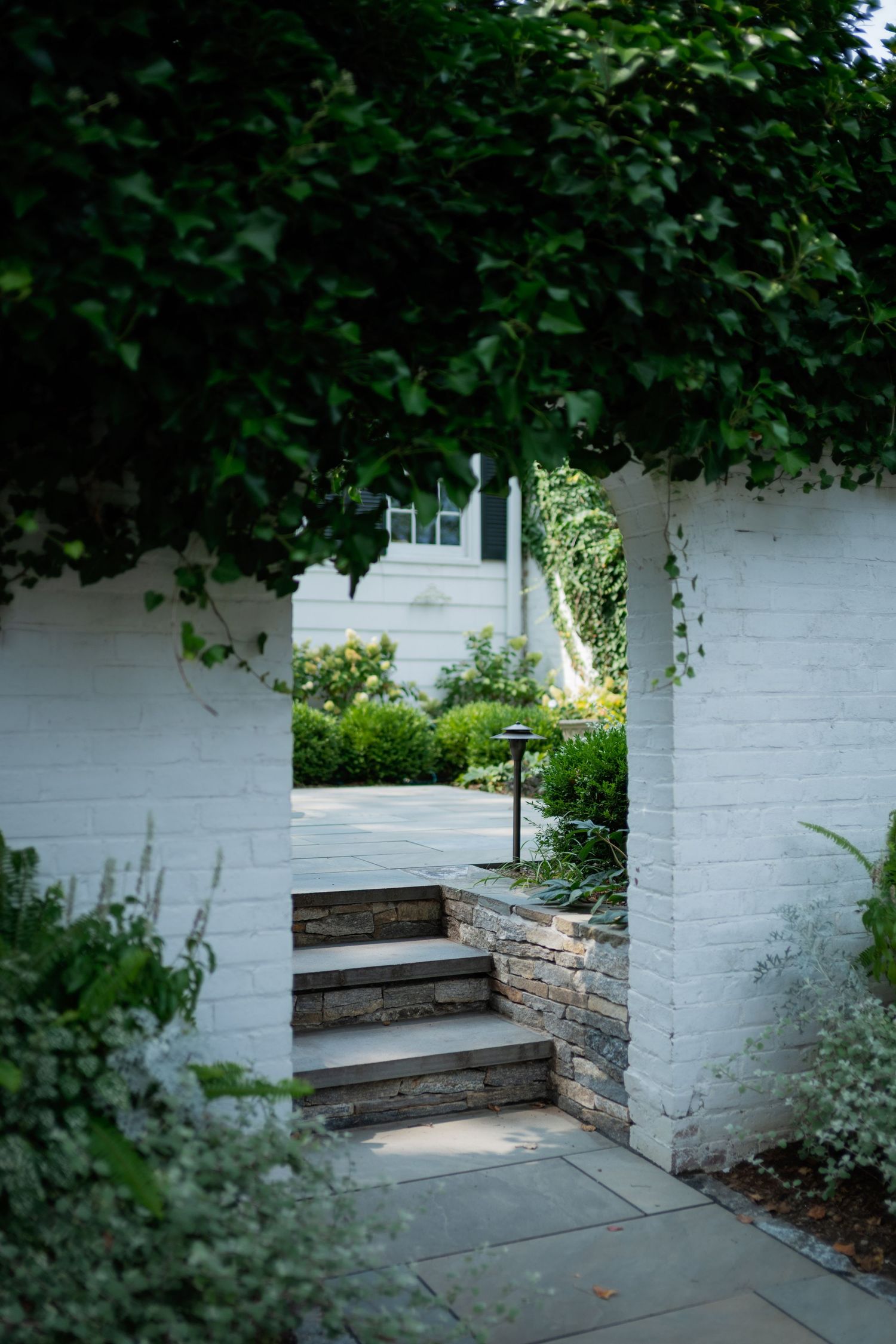 Stone steps with white painted brick walls lead to a garden path surrounded by lush green foliage.