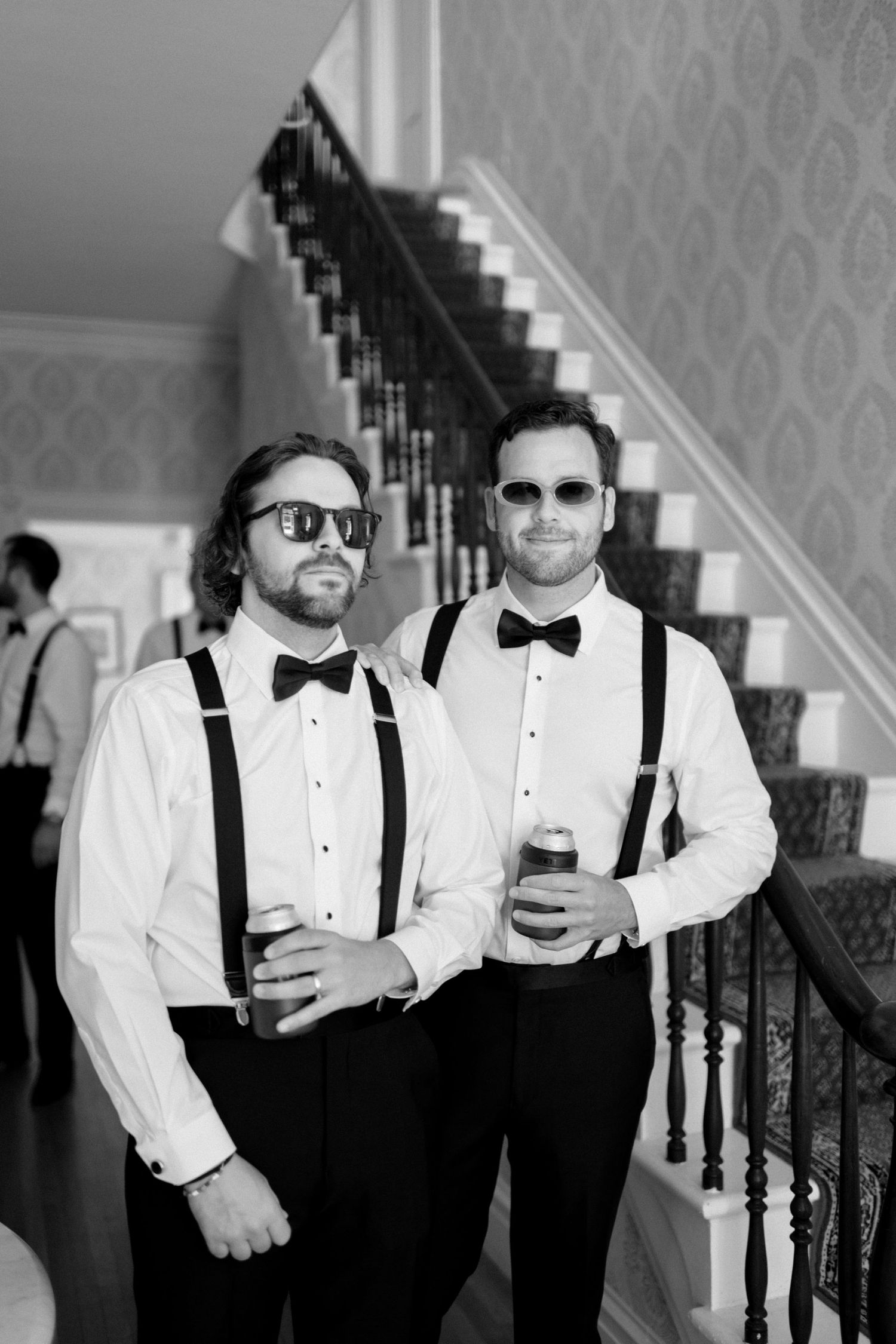 Two groomsmen in suspenders and bowties posing on staircase with drinks.