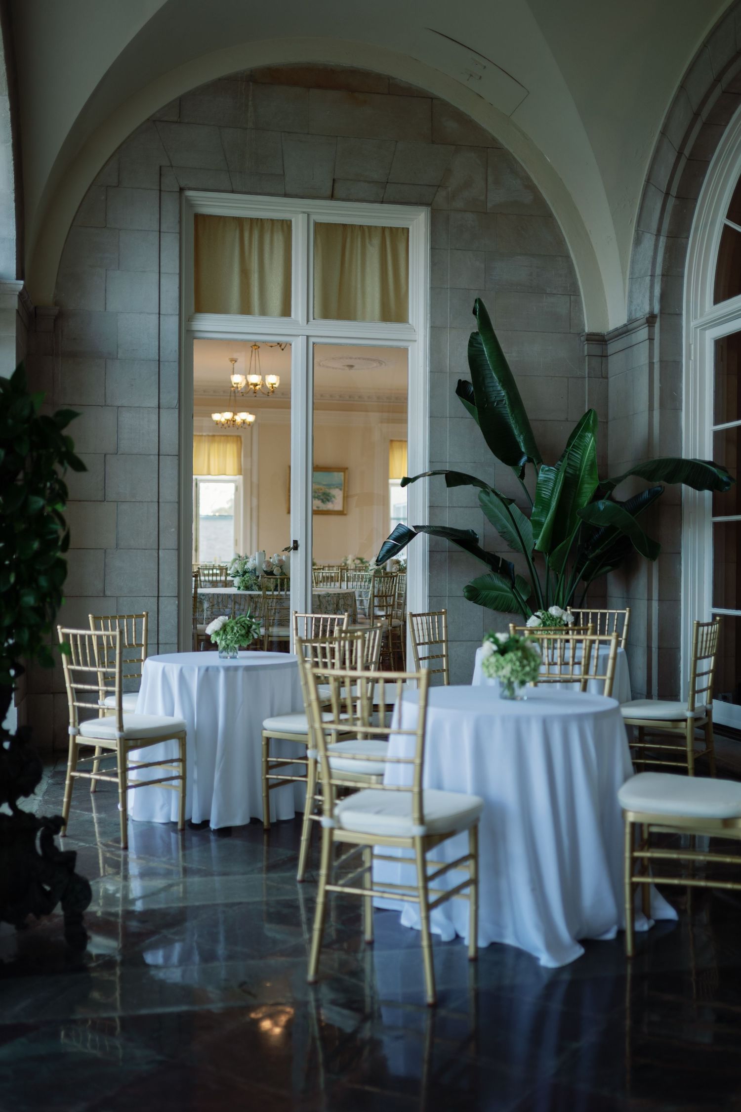 Elegant reception venue with white round tables, gold chairs, and arched windows in a classic stone interior.