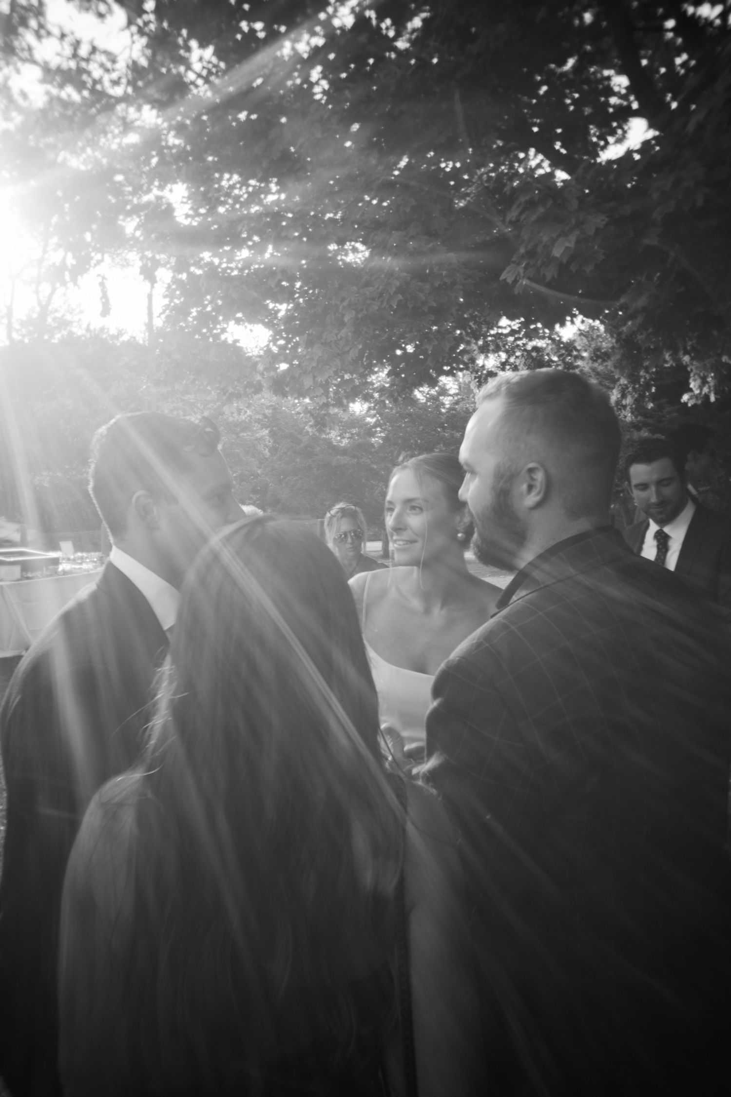 Black and white wedding photo of bride and groom sharing a moment outdoors with sun flare through trees.