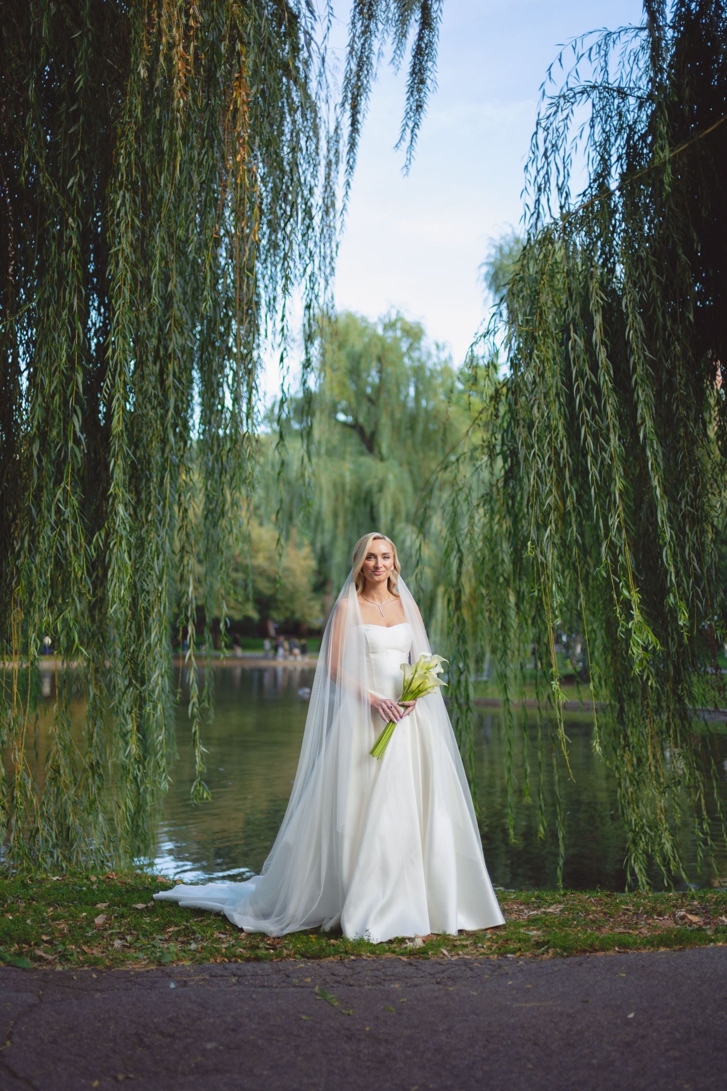 Bride in white gown and veil holding bouquet stands under weeping willow trees by a serene lake.