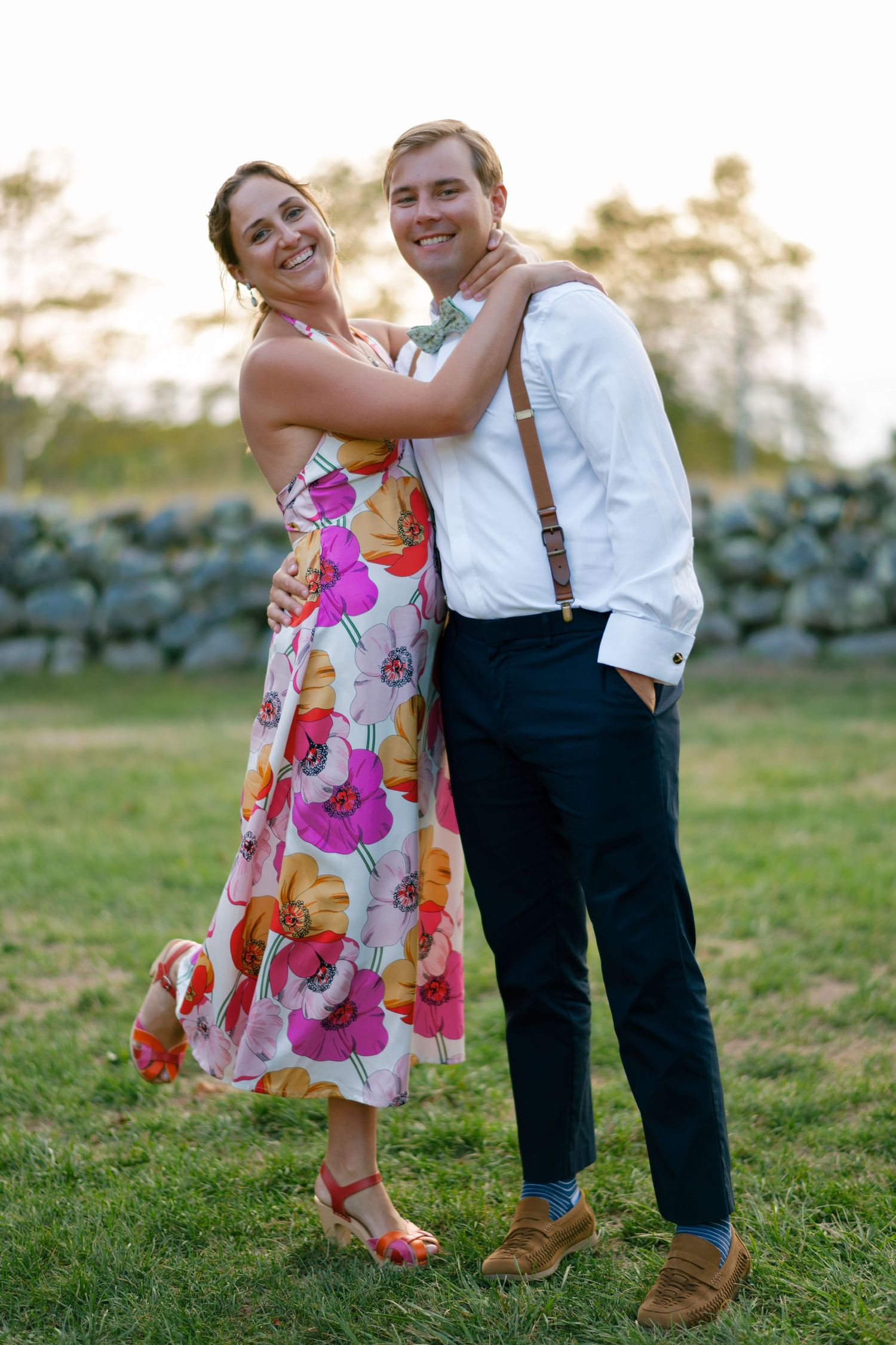 Smiling couple embracing outdoors, woman in floral dress, man in white shirt with suspenders, standing on green grass.