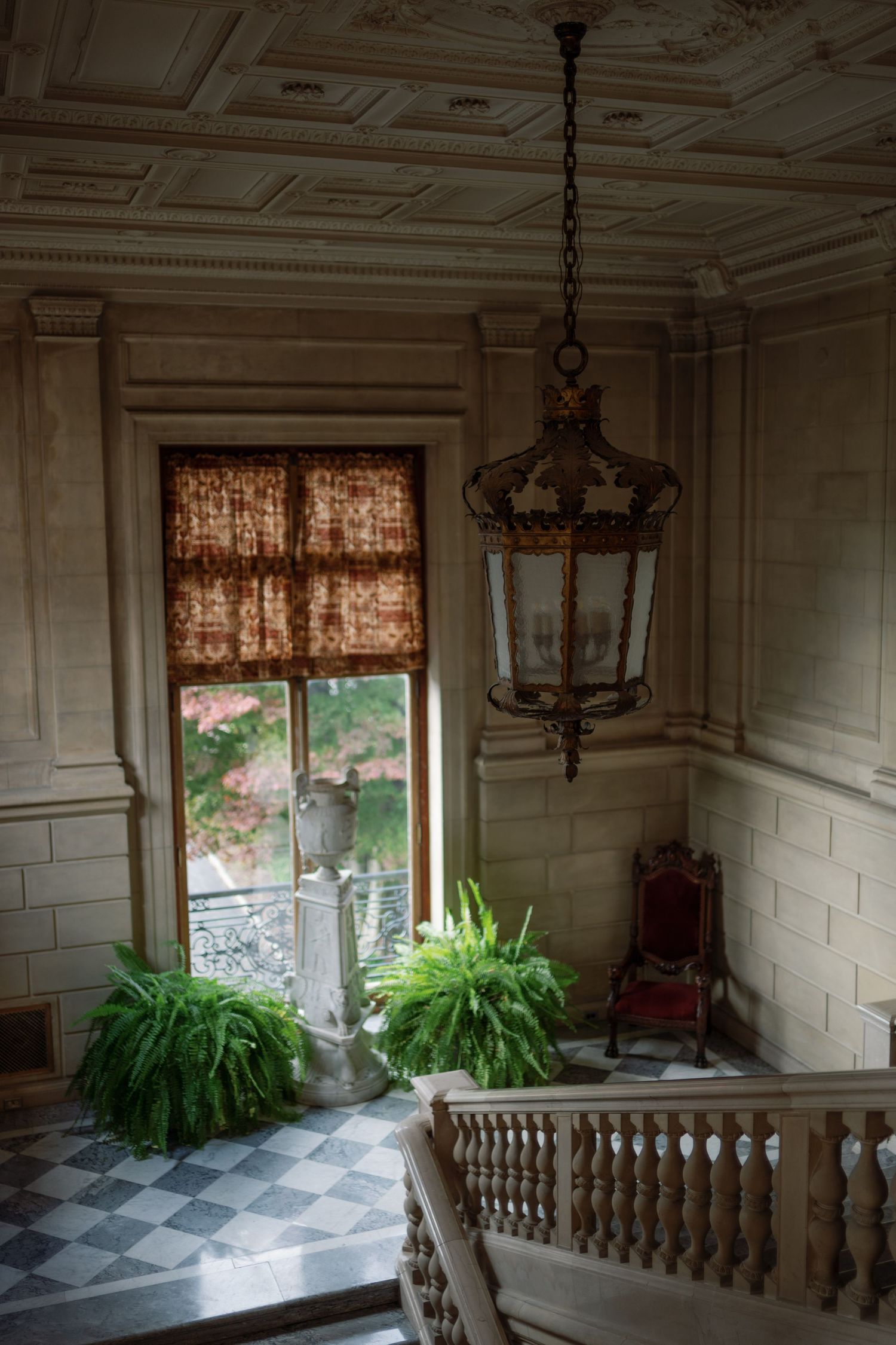Elegant historic interior with ornate ceiling, hanging chandeliers, white paneled walls, and lush green ferns by sunlit windows.