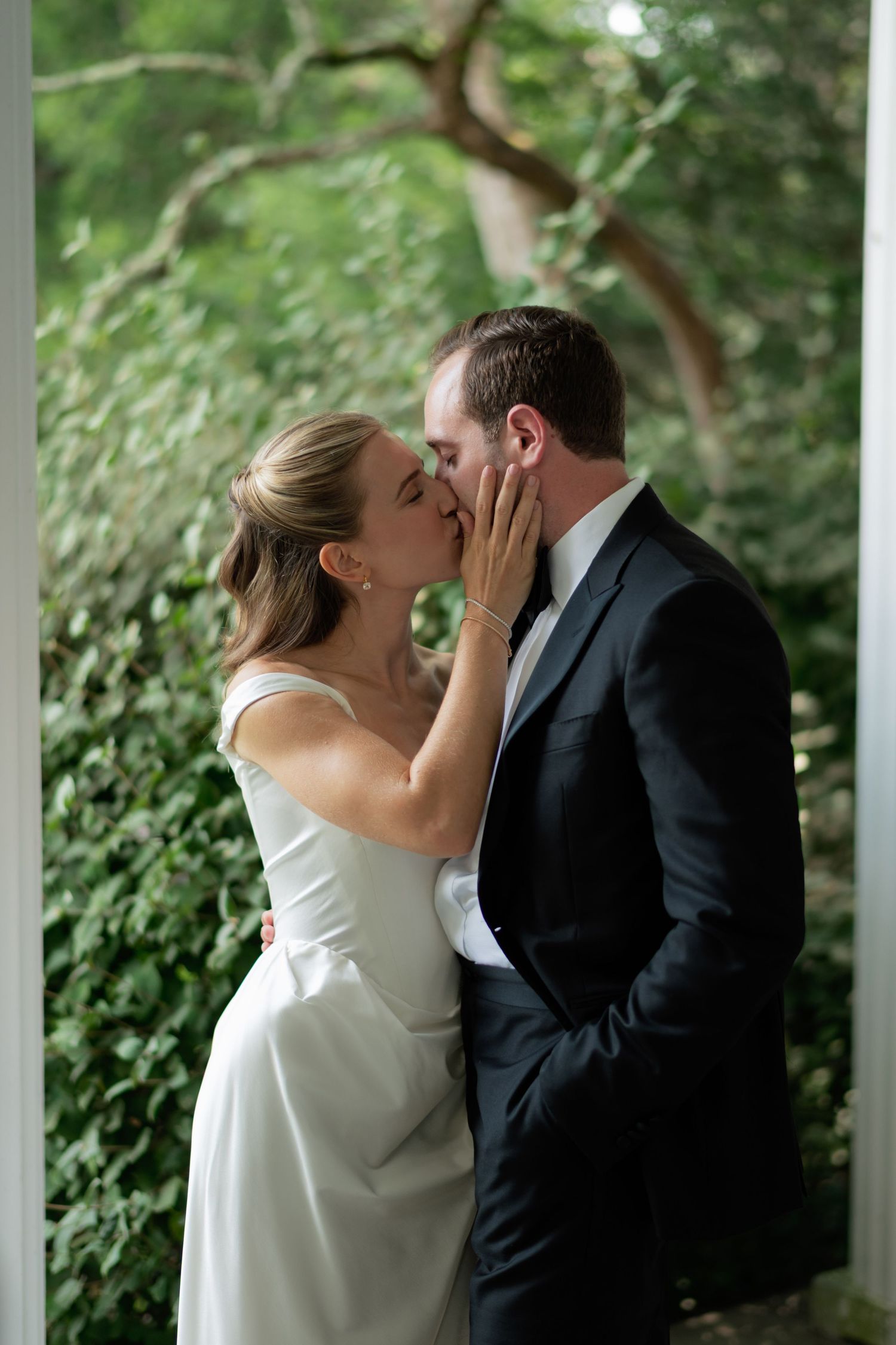 A romantic wedding couple shares an intimate kiss in front of a lush green garden backdrop.