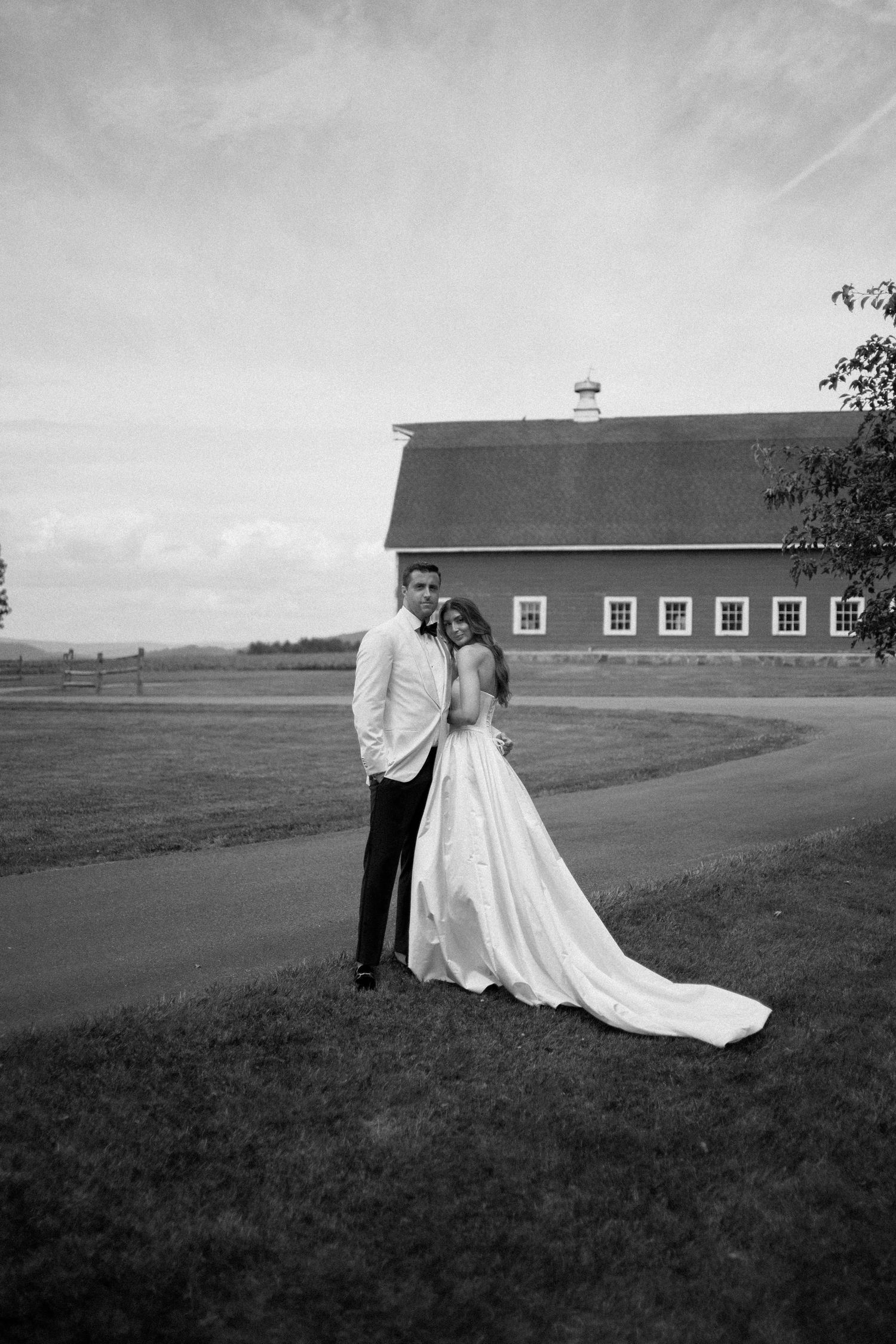 A series of black and white wedding photos showing a couple posing in front of a rustic barn on rolling hills.