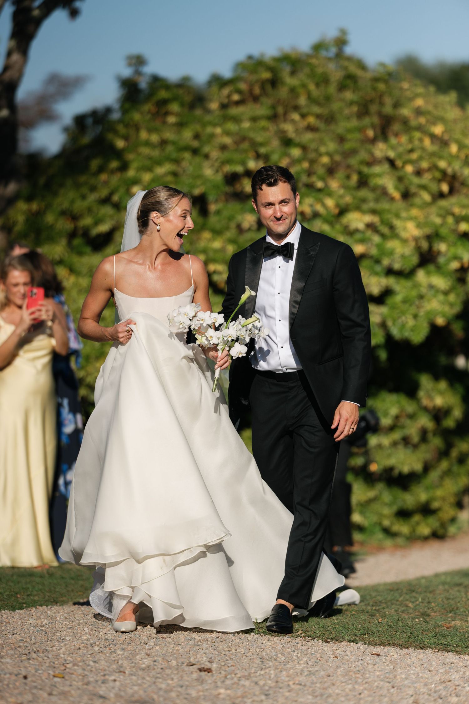 Joyful bride in white gown and groom in tuxedo walk together after wedding ceremony outdoors.