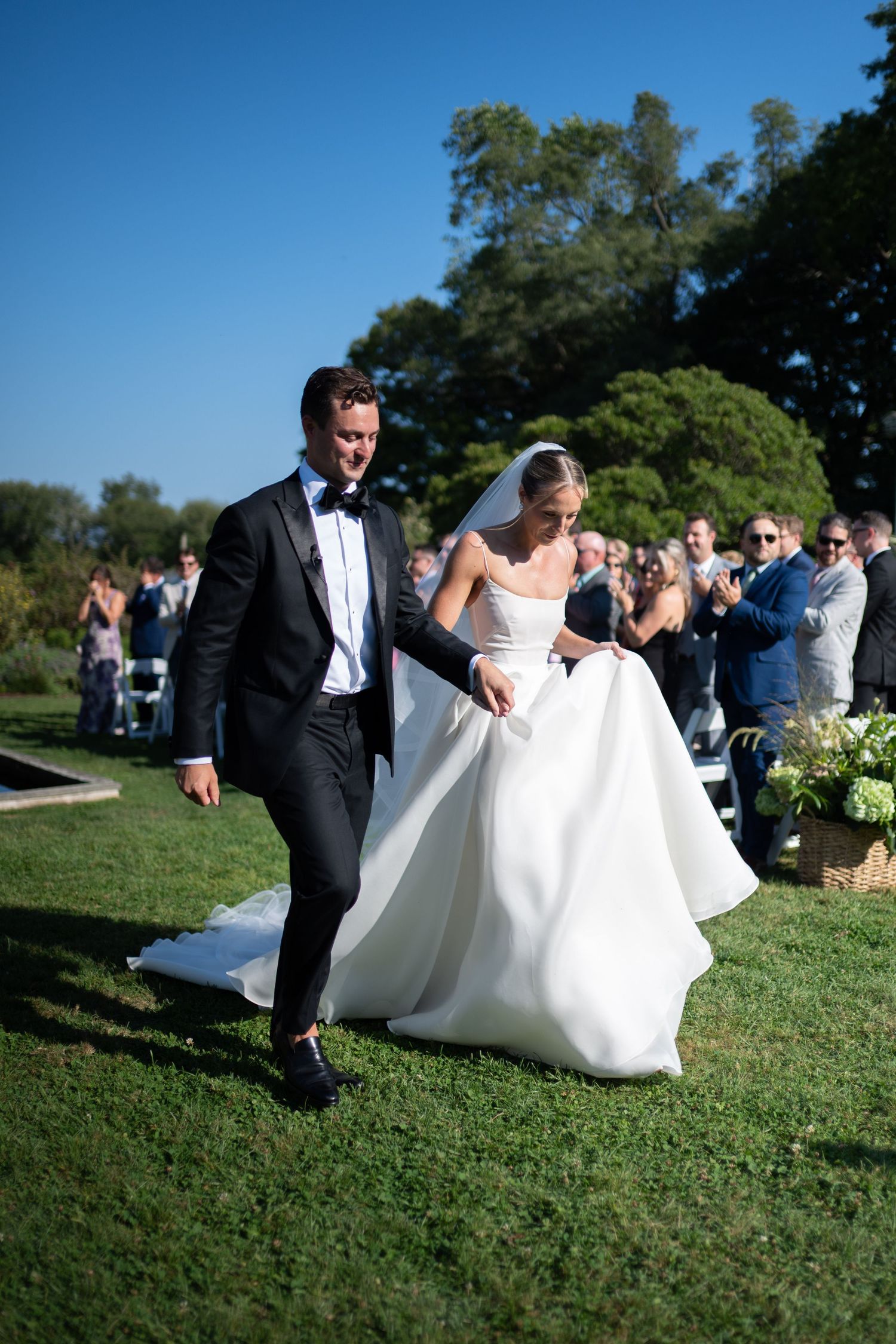 Bride and groom walk down outdoor wedding aisle, bride in white ball gown with veil, groom in black tuxedo.