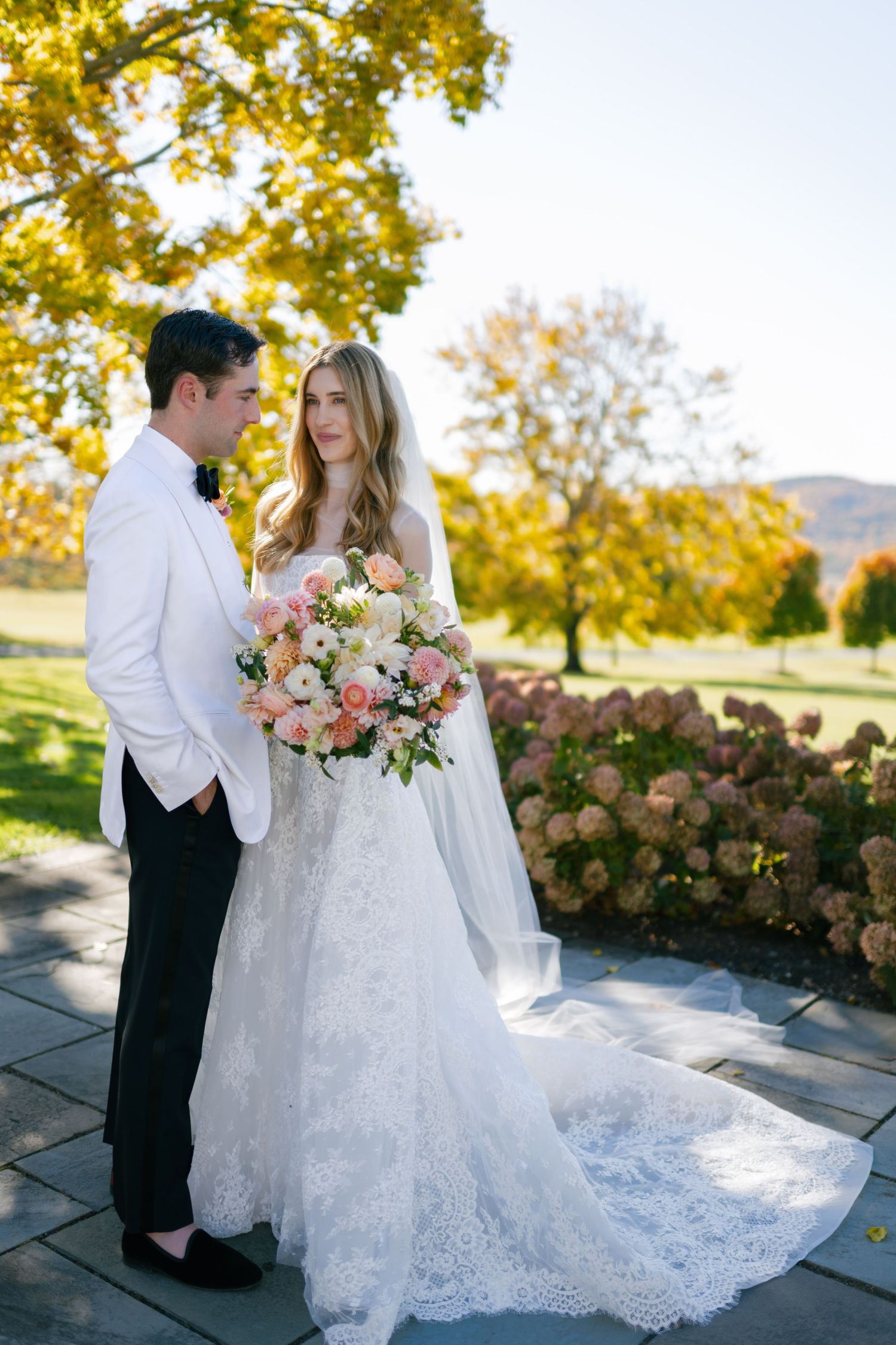 Couple in wedding attire posing outdoors with autumn foliage and a beautiful bouquet.
