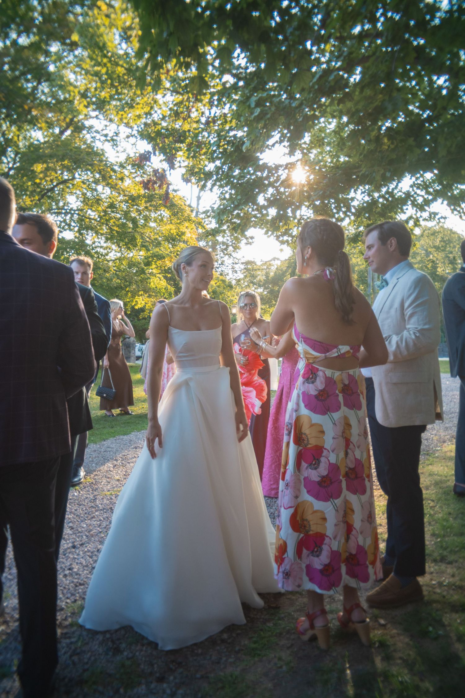 Bride in white gown chatting with guests in colorful dresses at an outdoor wedding reception under sunlit trees.