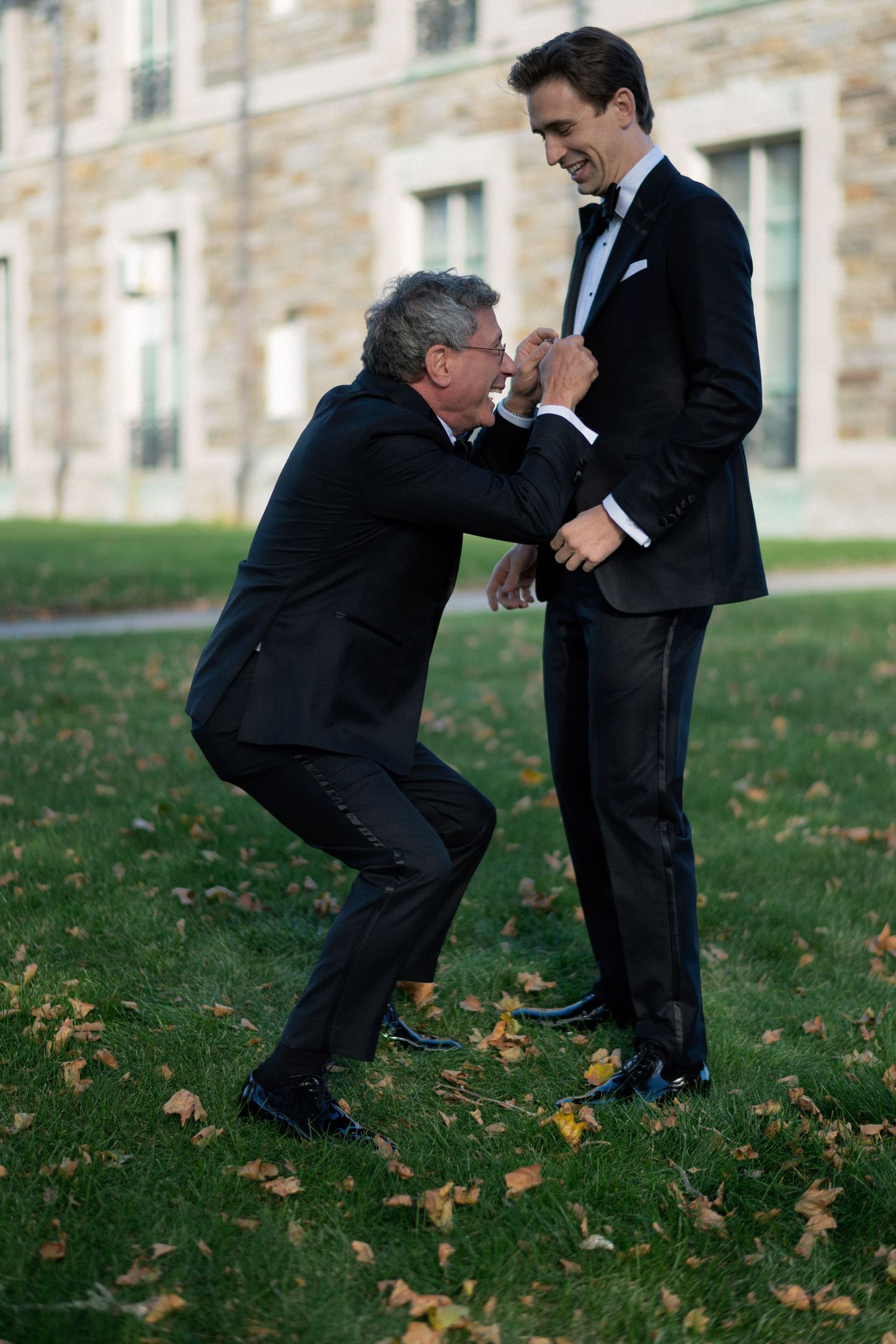 Two people in black suits playfully interact on a lawn with autumn leaves scattered around the stone building.