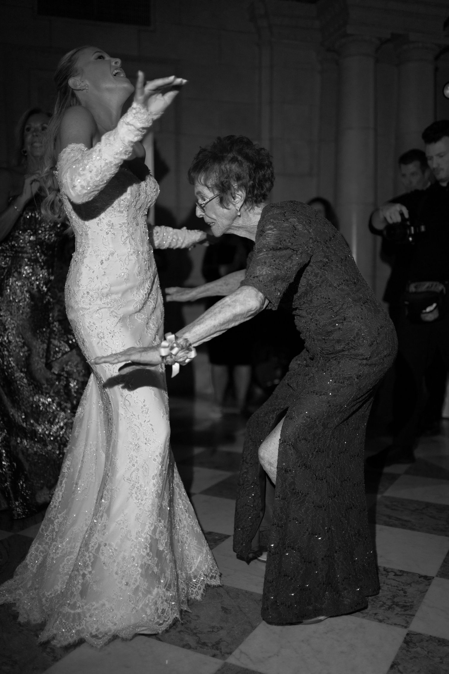 A bride in a white lace gown dances with an elderly guest in a dark dress at an elegant reception.
