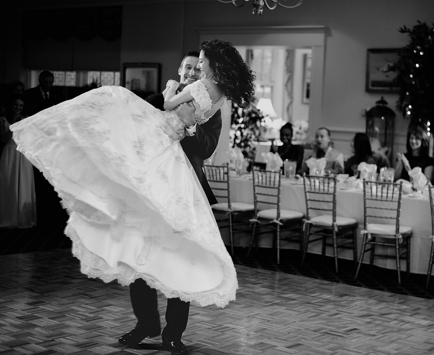 Black and white photo of groom spinning bride in wedding dress during first dance at elegant reception venue.