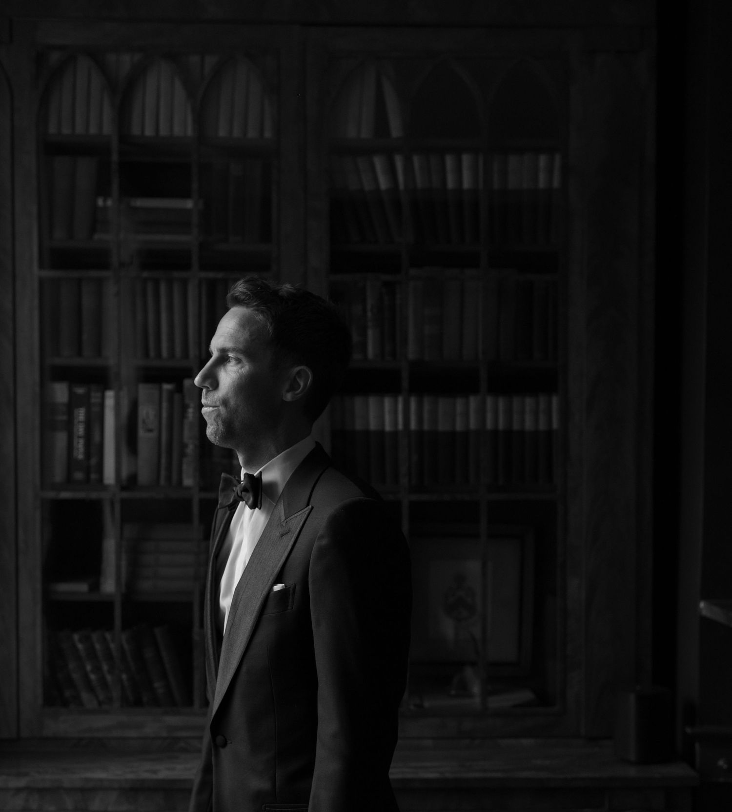 A formal portrait of a groom in a tuxedo and bowtie standing in a library.