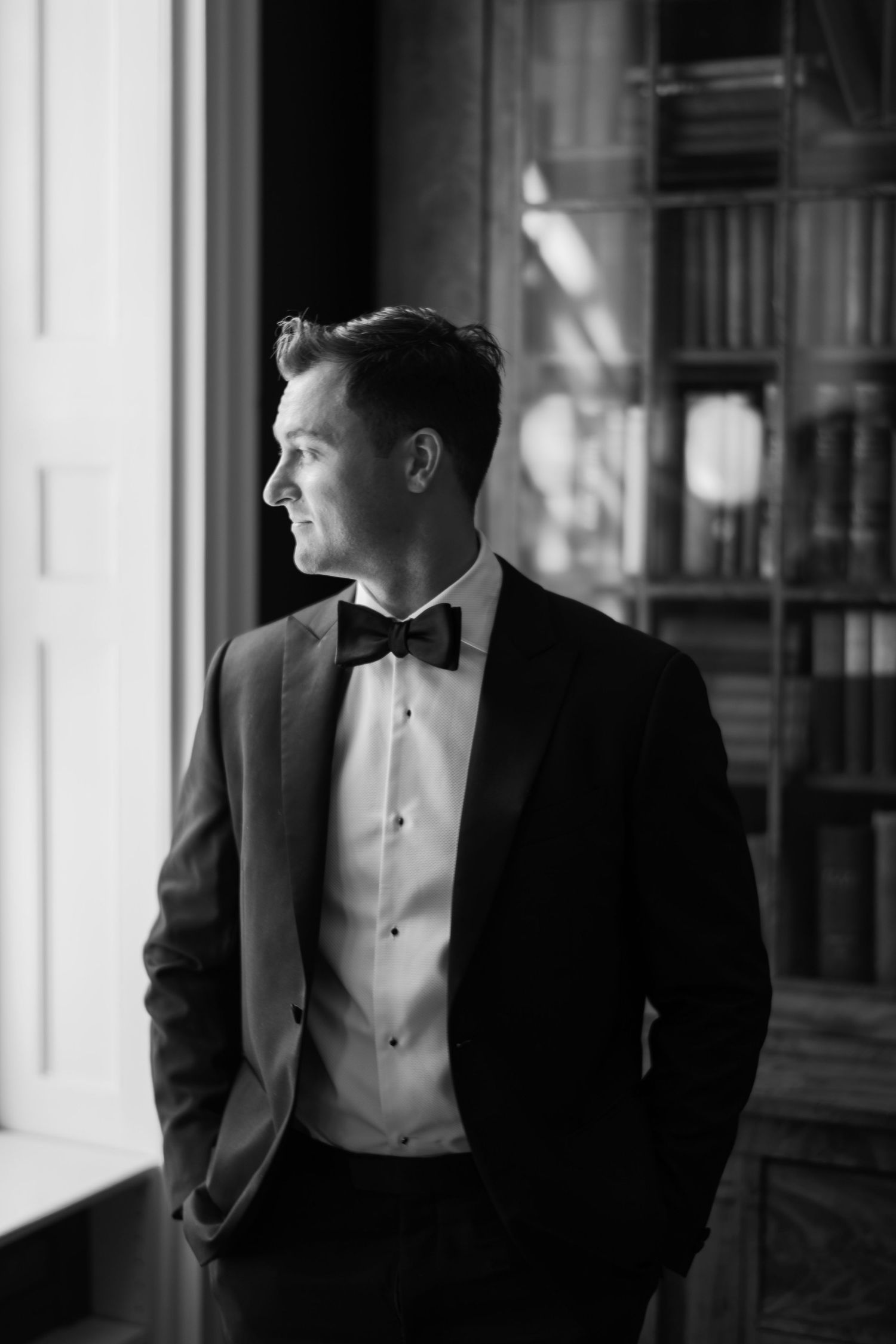 Black and white portrait of an elegant man in a tuxedo with bow tie standing by a window in a library.