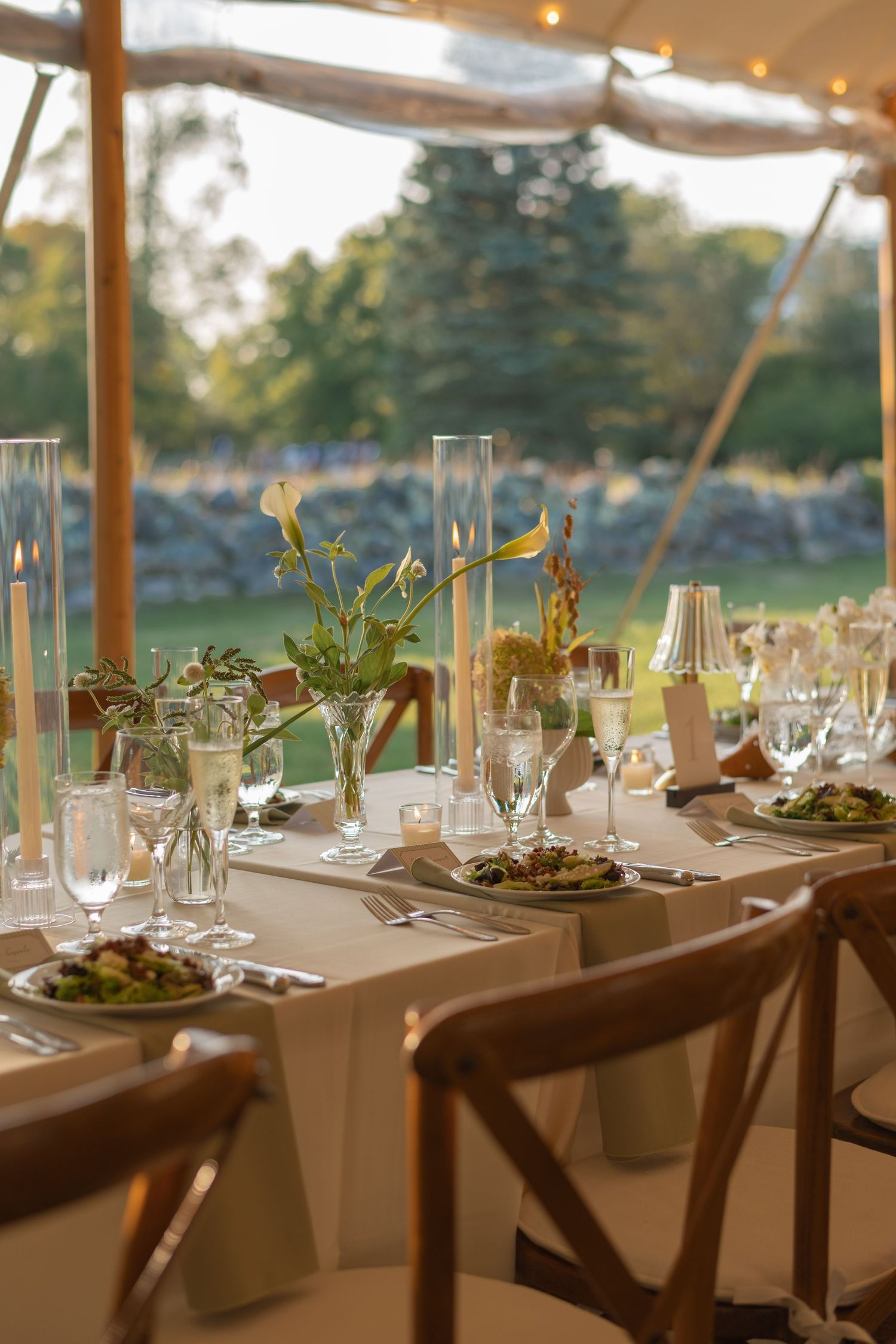 Elegant outdoor wedding reception table set with floral centerpieces, tall candles, and glassware under a tent.