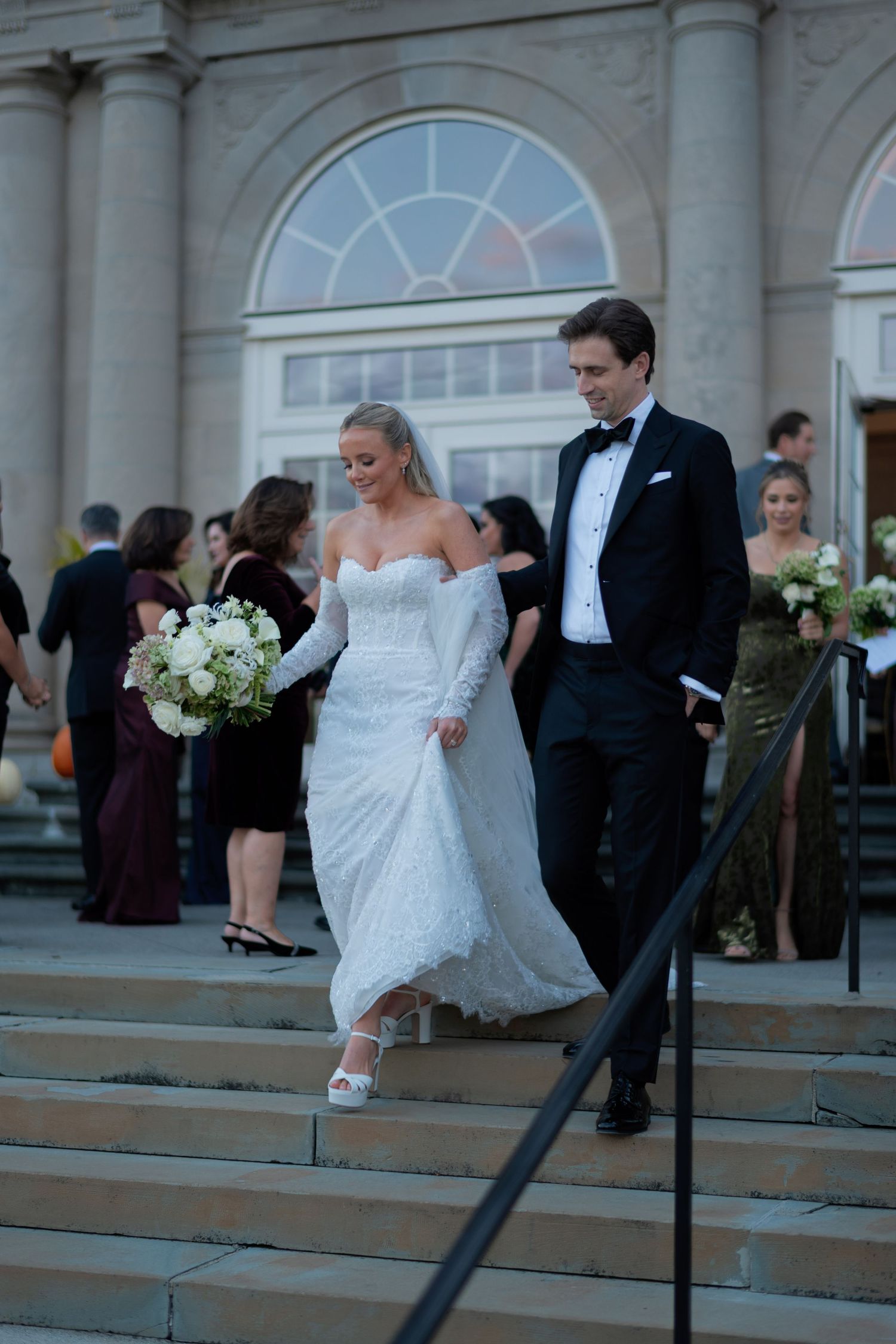 A wedding couple descends stone steps outside a grand building with elegant white columns and arched windows.
