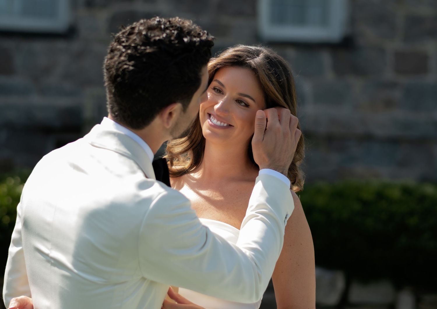 A groom in a white suit tenderly cups the face of a smiling bride outdoors at a wedding ceremony.
