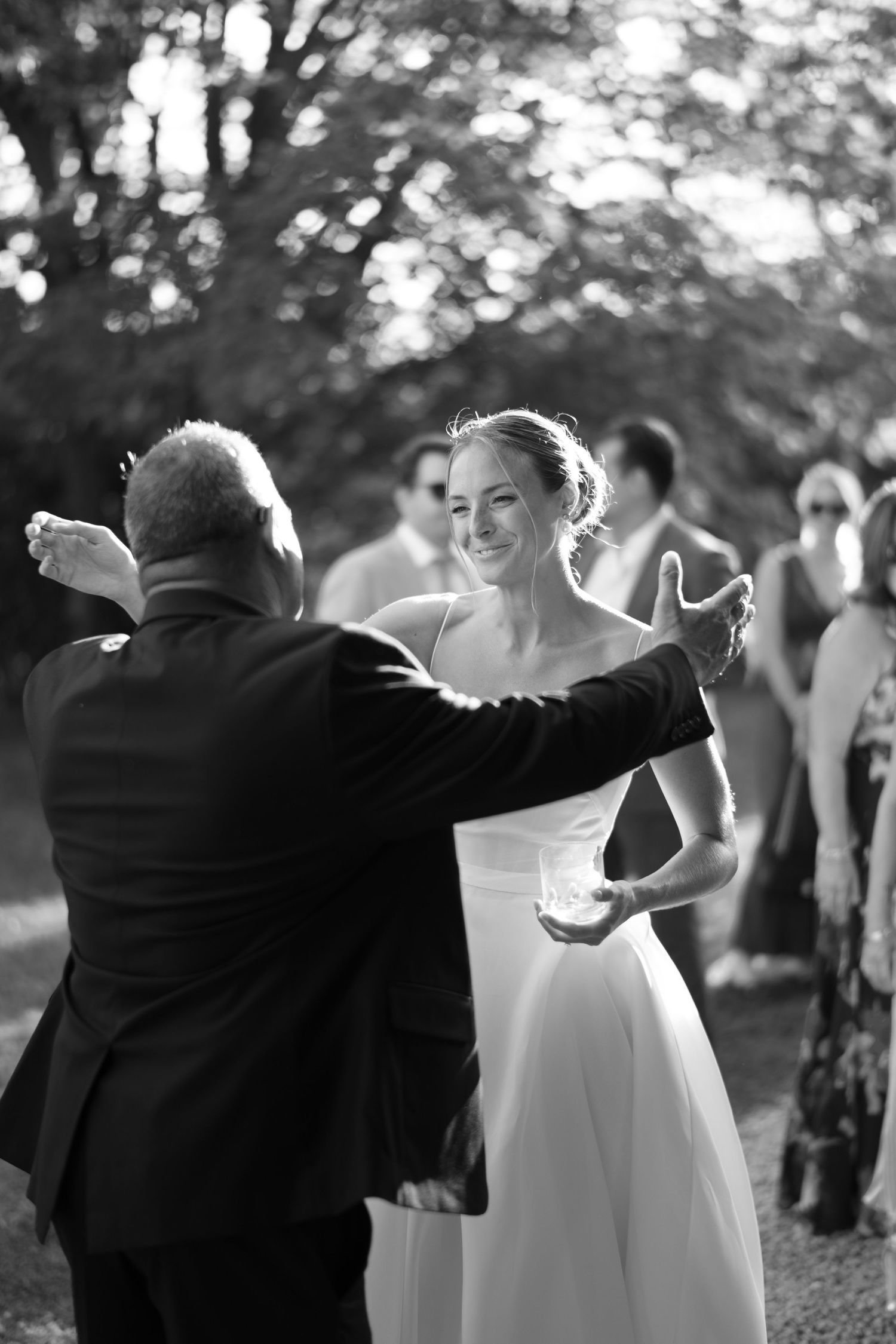 Black and white photo of a smiling bride dancing with a man in a suit at an outdoor wedding reception.