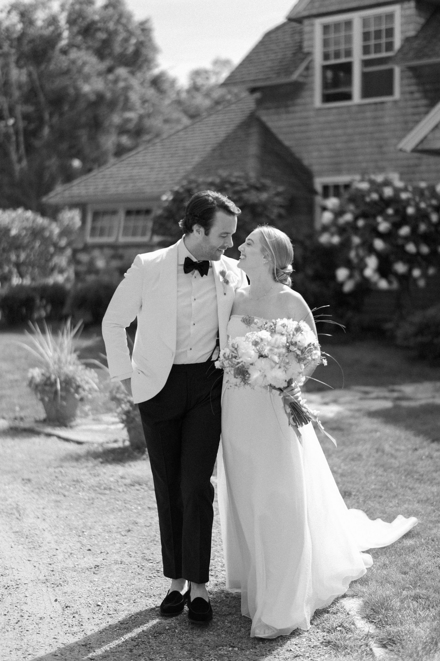 A couple in wedding attire share a moment outside a rustic house in black and white.