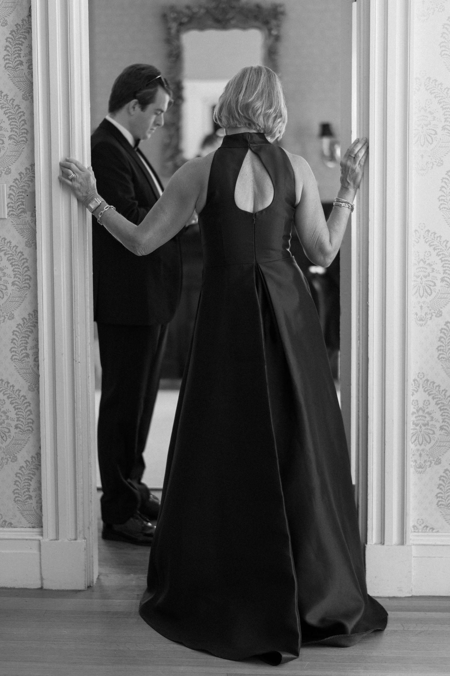 Formal couple in evening wear looking through doorway in black and white.