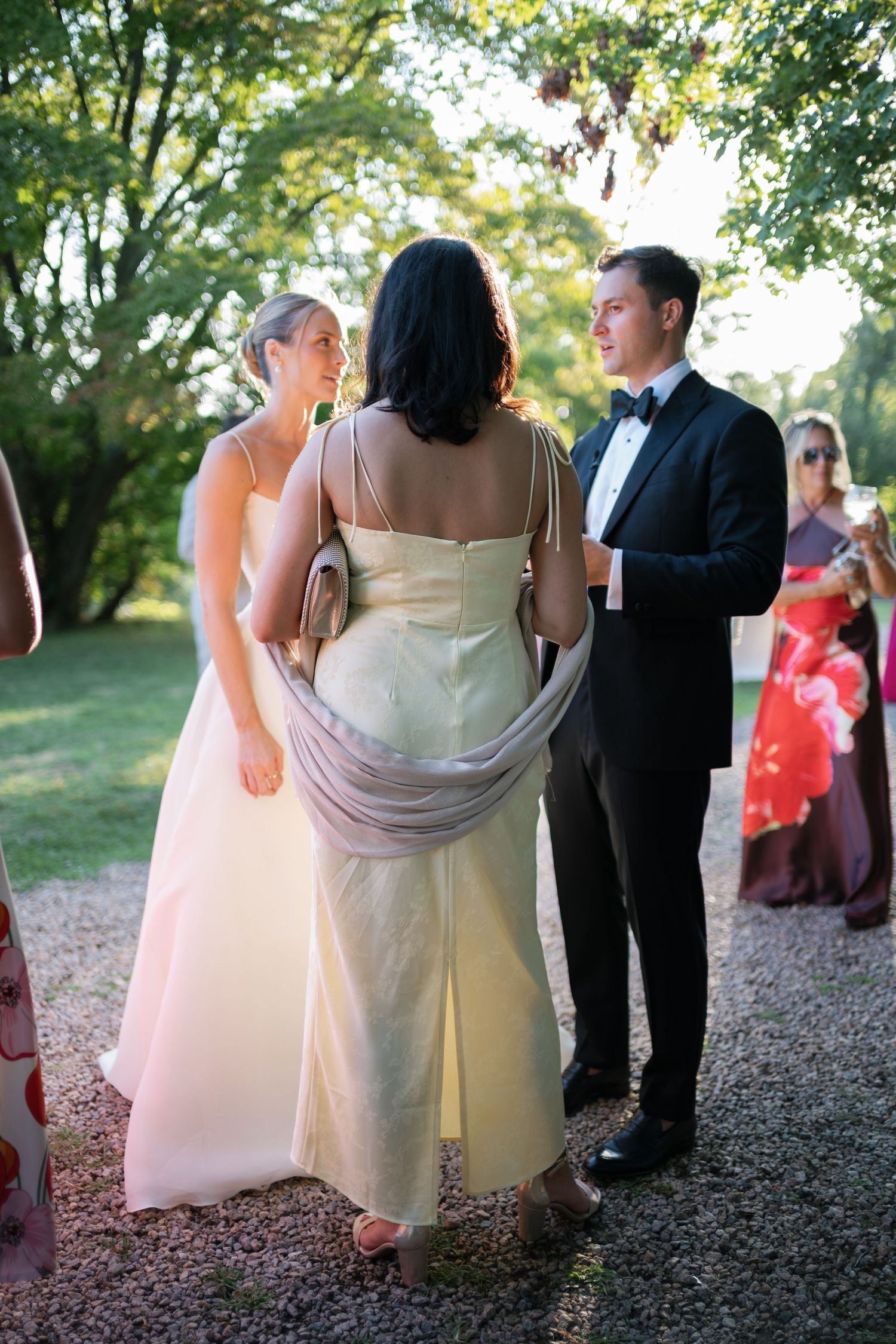 Wedding guests in formal attire converse outdoors, with bride in white gown and man in tuxedo on gravel path.