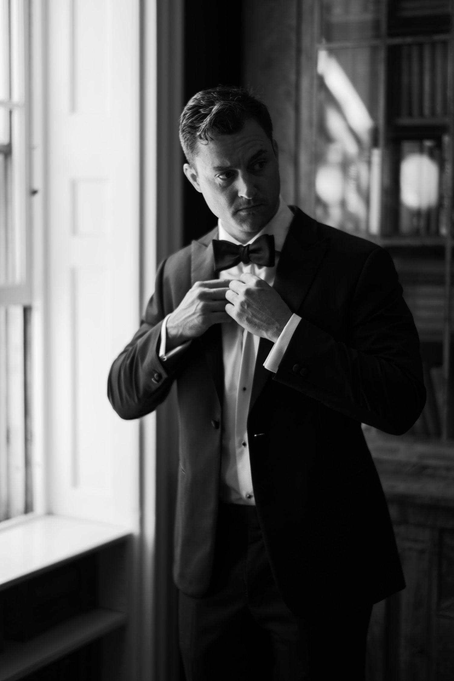 Black and white photo of a groom in a tuxedo adjusting his bow tie near a window.