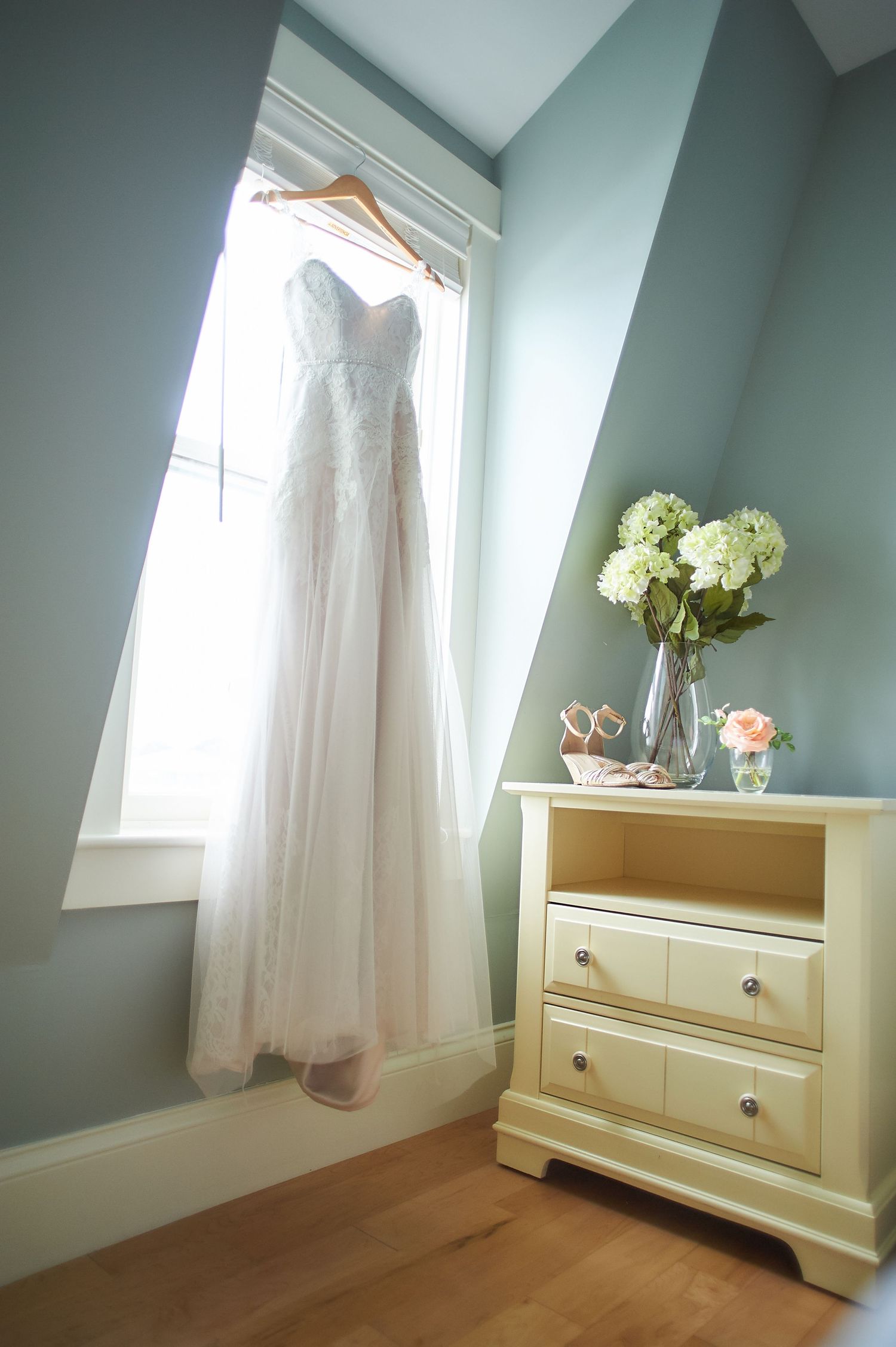 White wedding dress hanging by window with cream dresser and floral arrangement.
