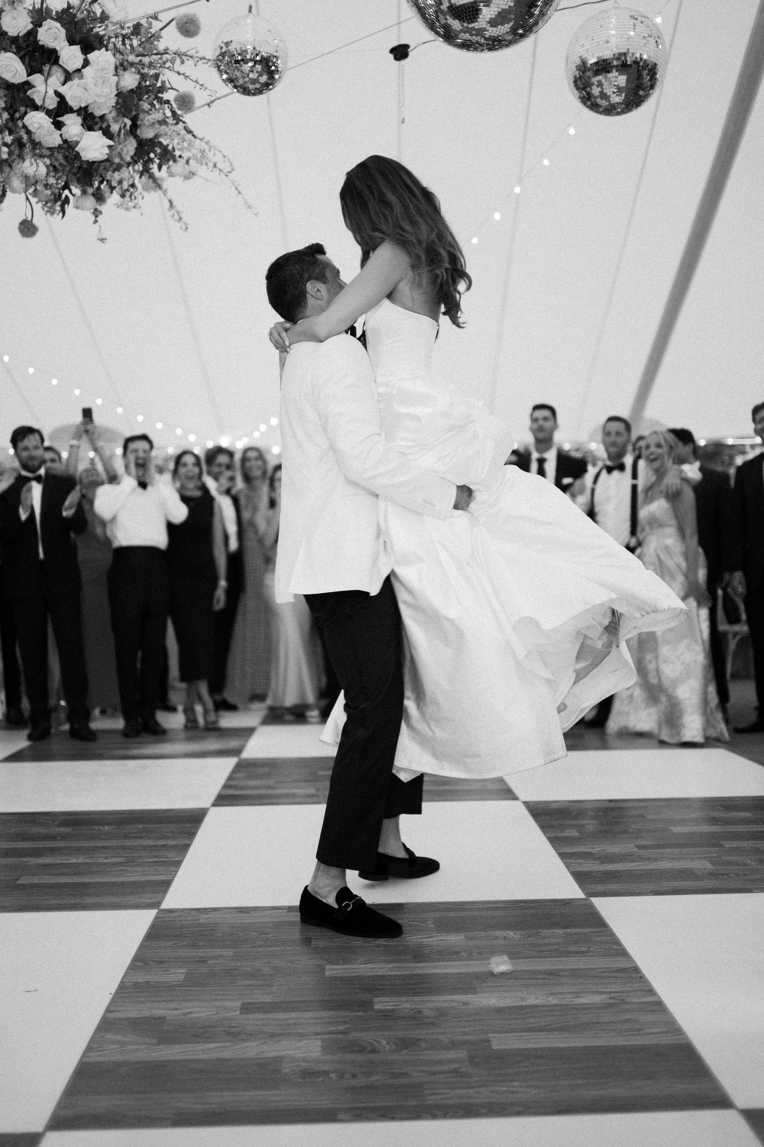 A couple shares their first dance at a wedding reception on a checkered dance floor under disco balls and floral decor.