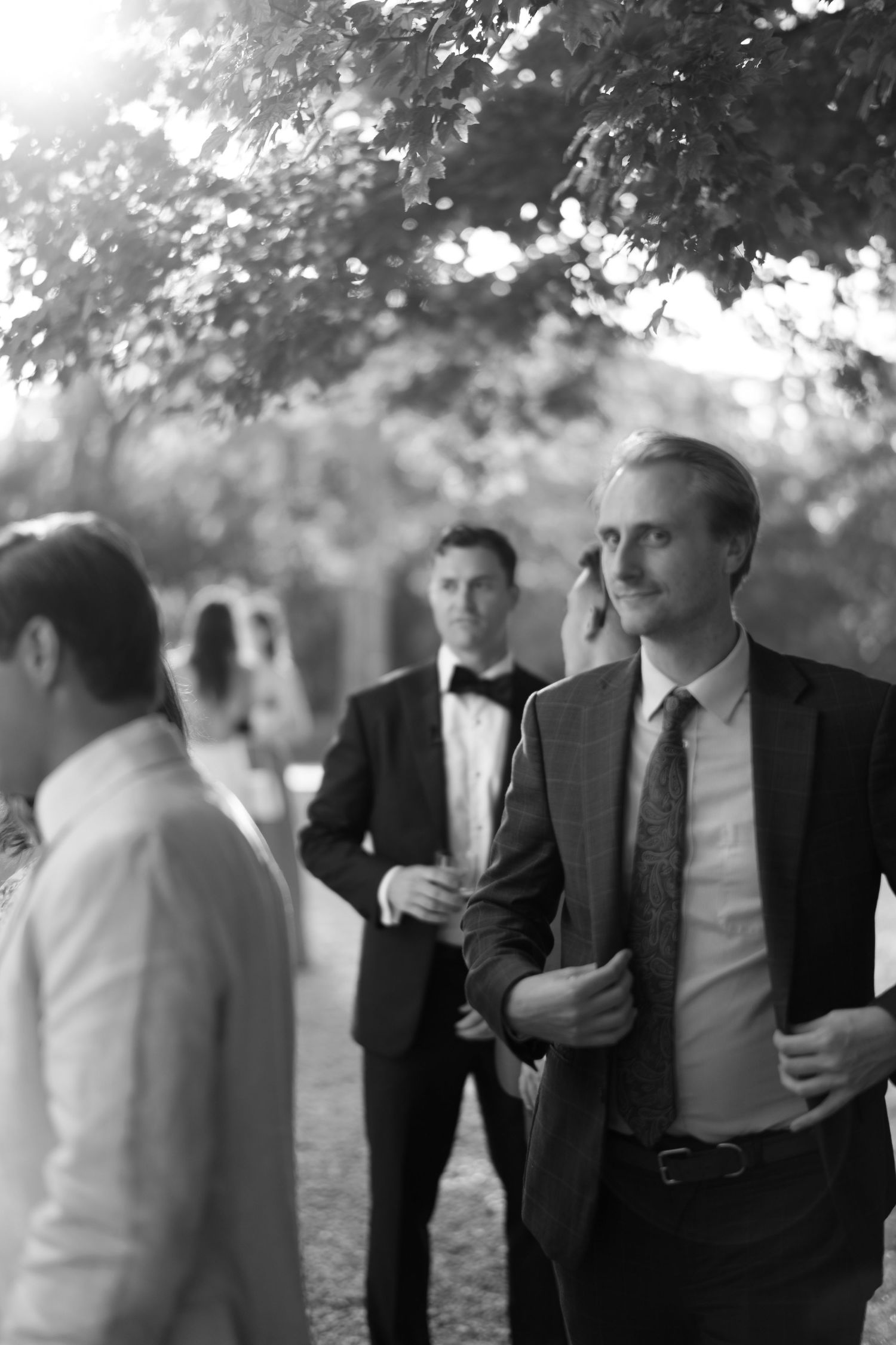 Black and white photo of elegantly dressed men in suits at a formal outdoor wedding event.