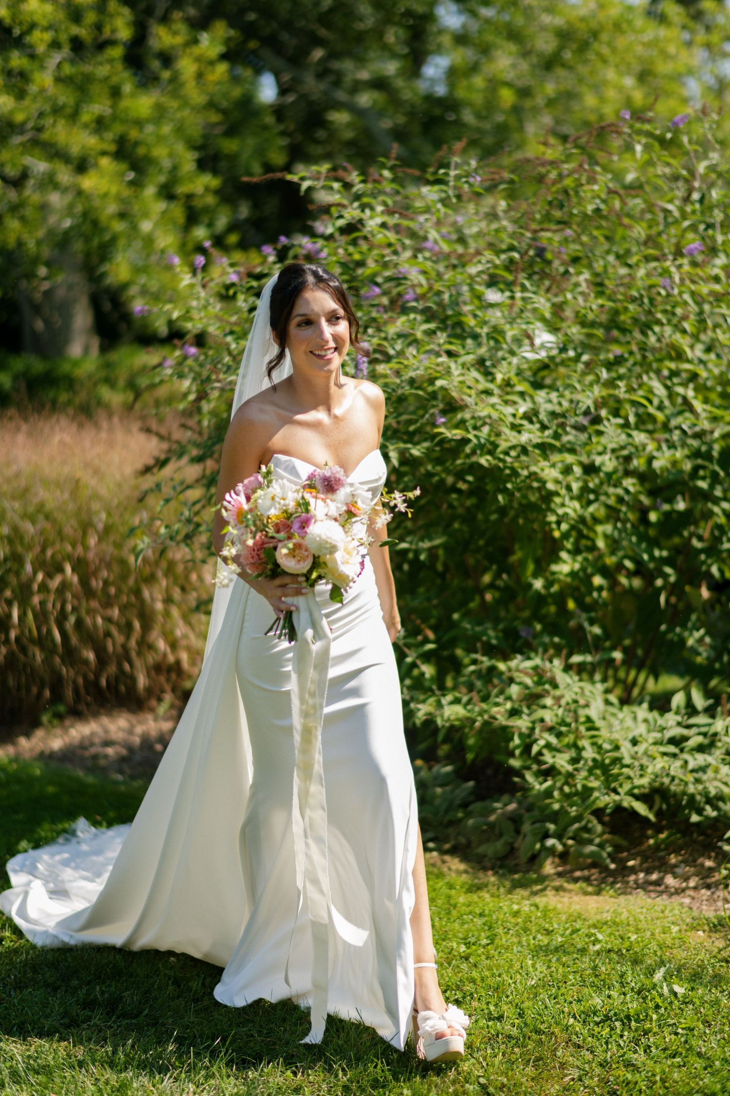 Elegant bride in off-shoulder white gown holding a colorful bouquet in a garden setting.